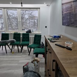 Oko - Pokój po remoncie z zielonymi krzesłami, niedokończone oświetlenie sufitowe i narzędzia na komodzie, widok na zimowe drzewa za oknem.