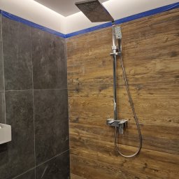 Oko - Nowoczesna kabina prysznicowa z deszczownicą, wykończona płytkami imitującymi drewno oraz ciemnymi płytkami kamiennymi. Niebieska taśma zabezpieczająca widoczna na górze ściany.