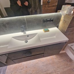 Oko - Nowoczesna umywalka z szarą szafką, drewnopodobne płytki podłogowe, odbicie osoby z telefonem w lustrze, widoczne gniazdka elektryczne nad umywalką.