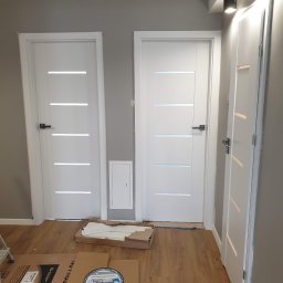 Oko - Montaż dwóch białych drzwi wewnętrznych z poziomymi przeszkleniami, widoczna puszka z farbą podkładową Reflex Grunt i kartony ochronne na podłodze.