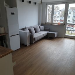 Oko - Jasne wnętrze pokoju z szarą sofą z poduszkami, białą lodówką, drewnianą podłogą i dużym oknem balkonowym z widokiem na budynki na zewnątrz.