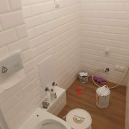 Oko - Biała łazienka w trakcie prac wykończeniowych, widoczne białe płytki typu metro, niedokończona toaleta, wiadra z farbą Beckers i Inisz oraz narzędzia glazurnicze na podłodze imitującej drewno.