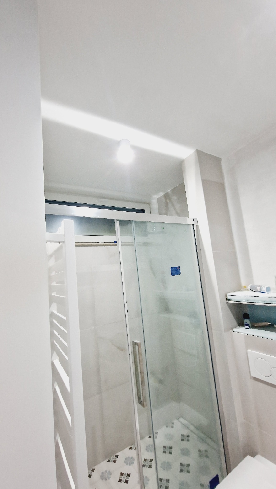 Biała kabina prysznicowa z przesuwanymi drzwiami i grzejnikiem drabinkowym w odnowionej łazience, widok na wzorzystą podłogę i półki z kosmetykami.