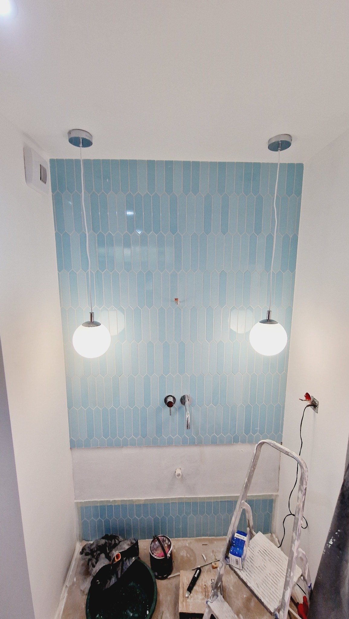Niebieskie, podłużne płytki ułożone pionowo na ścianie łazienki, widoczne chromowane krany, wiszące lampy kuliste, drabina i narzędzia na podłodze w trakcie prac wykończeniowych.