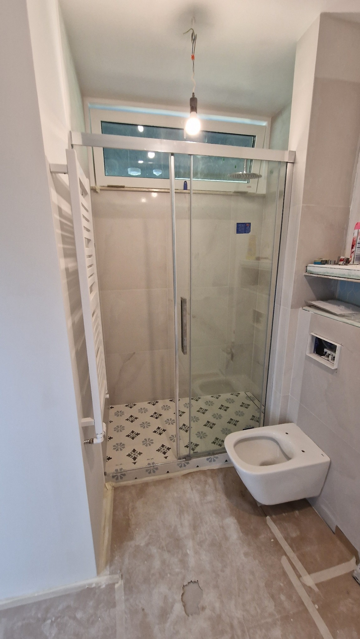 Niedokończony remont łazienki: kabina prysznicowa z przesuwnymi drzwiami, wisząca toaleta, grzejnik drabinkowy, dekoracyjne płytki pod prysznicem, surowa podłoga w trakcie prac.