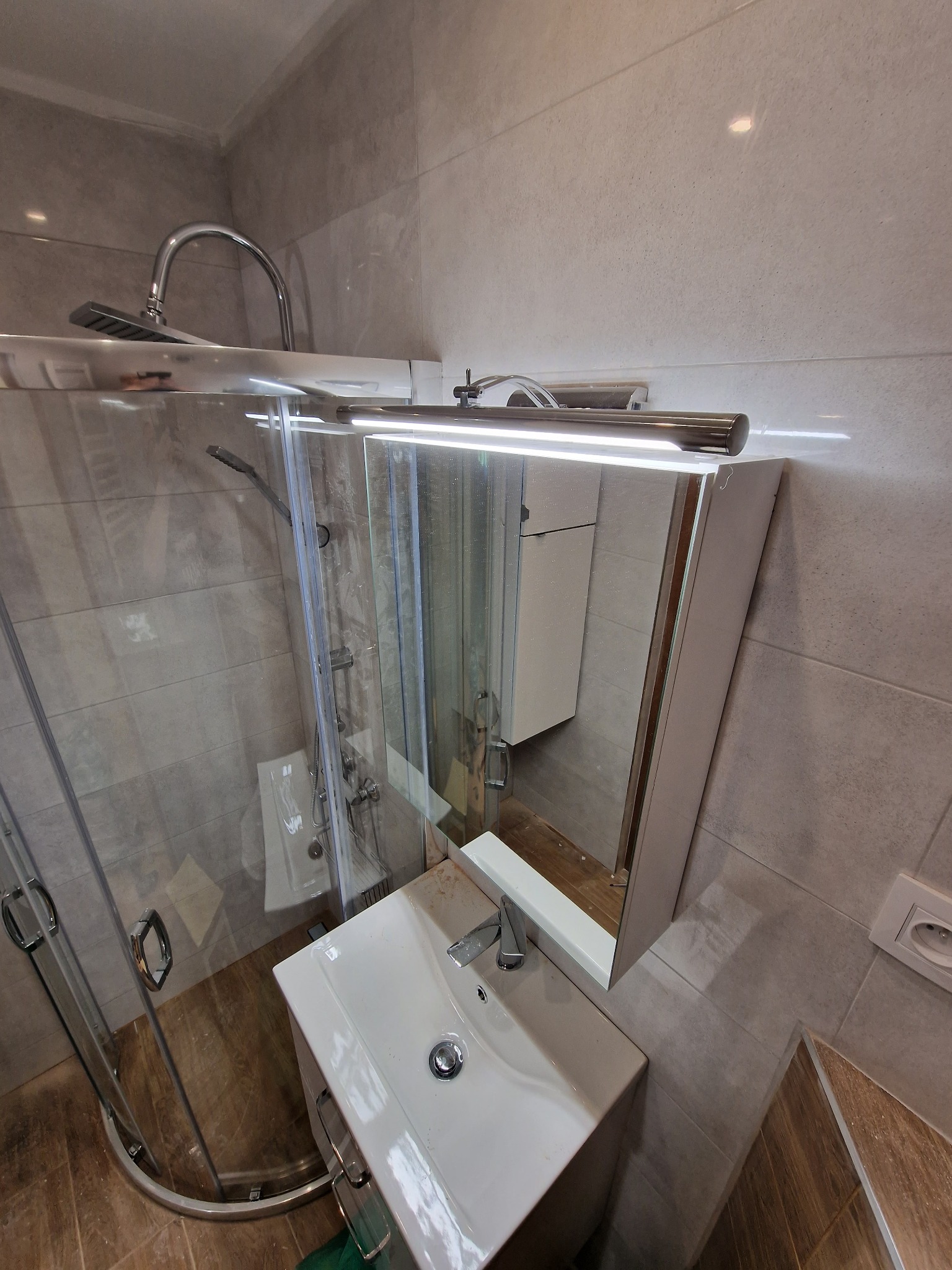 Nowoczesna łazienka z kabiną prysznicową z zaokrąglonymi drzwiami, umywalką z szafką i lustrem z oświetleniem LED, wykończona płytkami w odcieniach szarości i brązu.