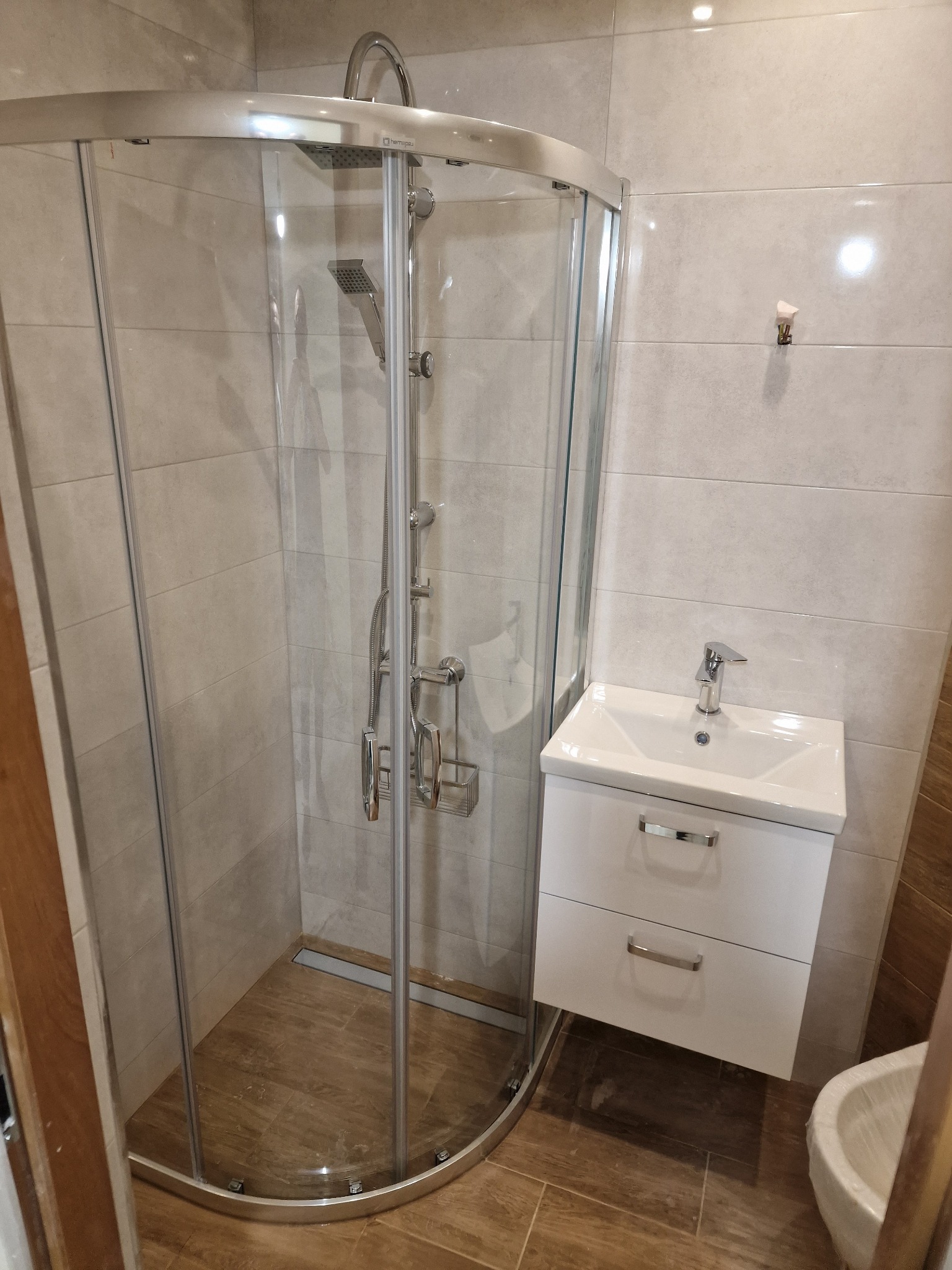 Nowoczesna łazienka po remoncie: kabina prysznicowa z chromowanymi detalami, biała szafka pod umywalką z dwiema szufladami i bateria umywalkowa.