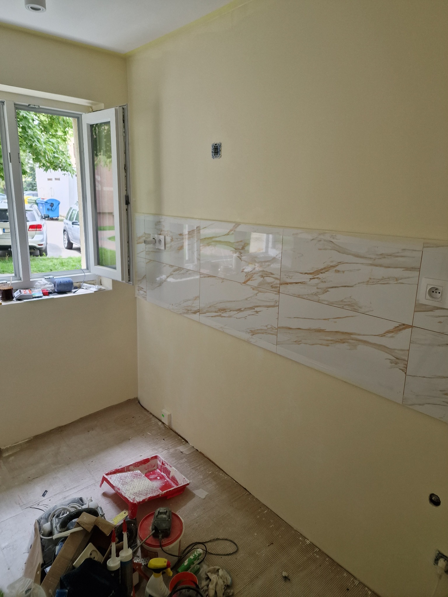 Fragment ściany w trakcie układania płytek imitujących marmur z widocznymi gniazdkami elektrycznymi, okno z widokiem na zewnątrz i nieporządek z narzędziami na podłodze.