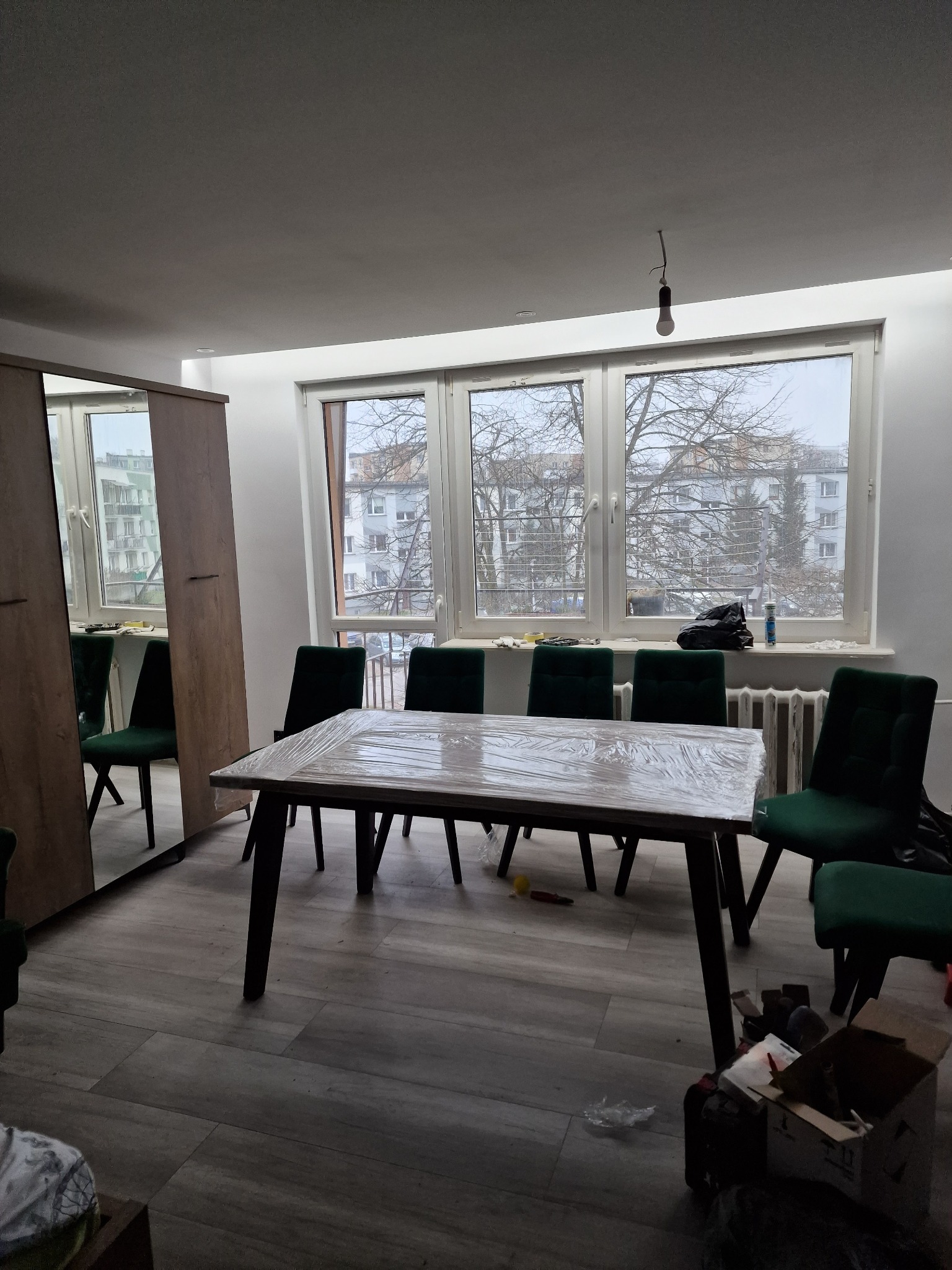 Pokój po remoncie z nową podłogą, stołem zabezpieczonym folią i zielonymi krzesłami. Widoczna szafa z lustrem i okno z widokiem na budynki.