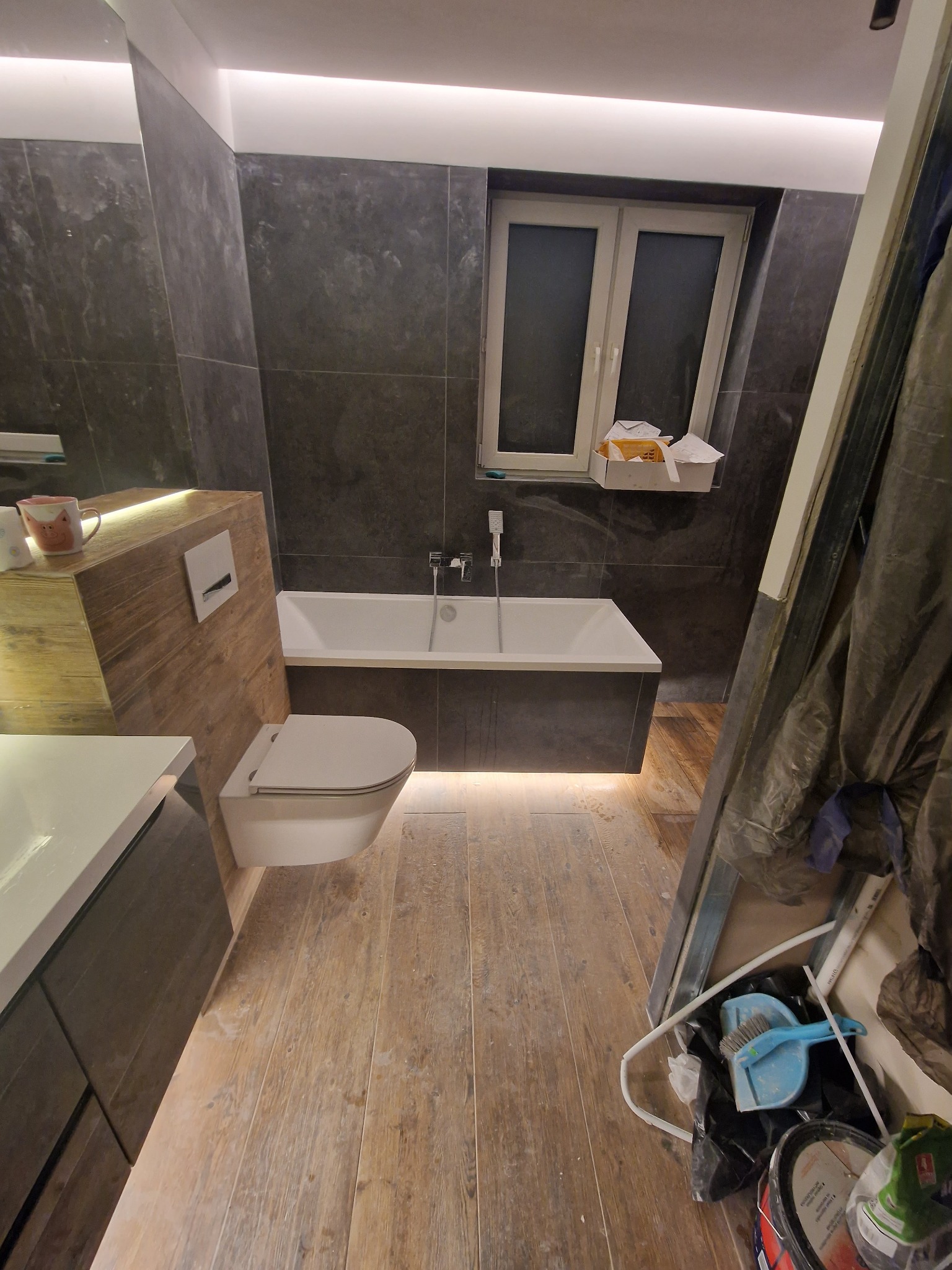 Nowoczesna łazienka w trakcie wykańczania, z podświetlaną wanną, wiszącą toaletą i drewnopodobną zabudową umywalki, widoczne narzędzia i materiały budowlane.