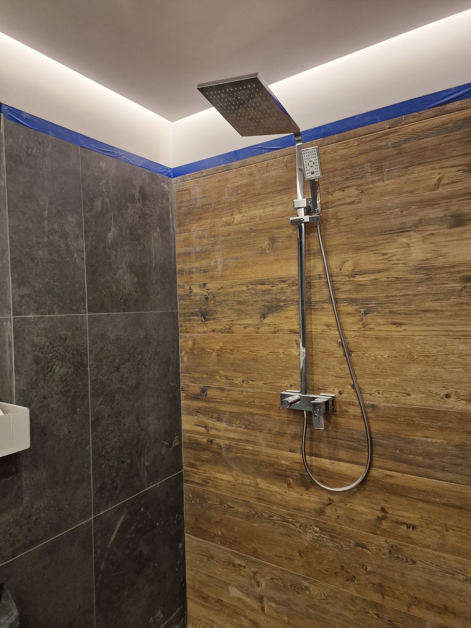 Nowoczesna kabina prysznicowa z deszczownicą, wykończona płytkami imitującymi drewno oraz ciemnymi płytkami kamiennymi. Niebieska taśma zabezpieczająca widoczna na górze ściany.