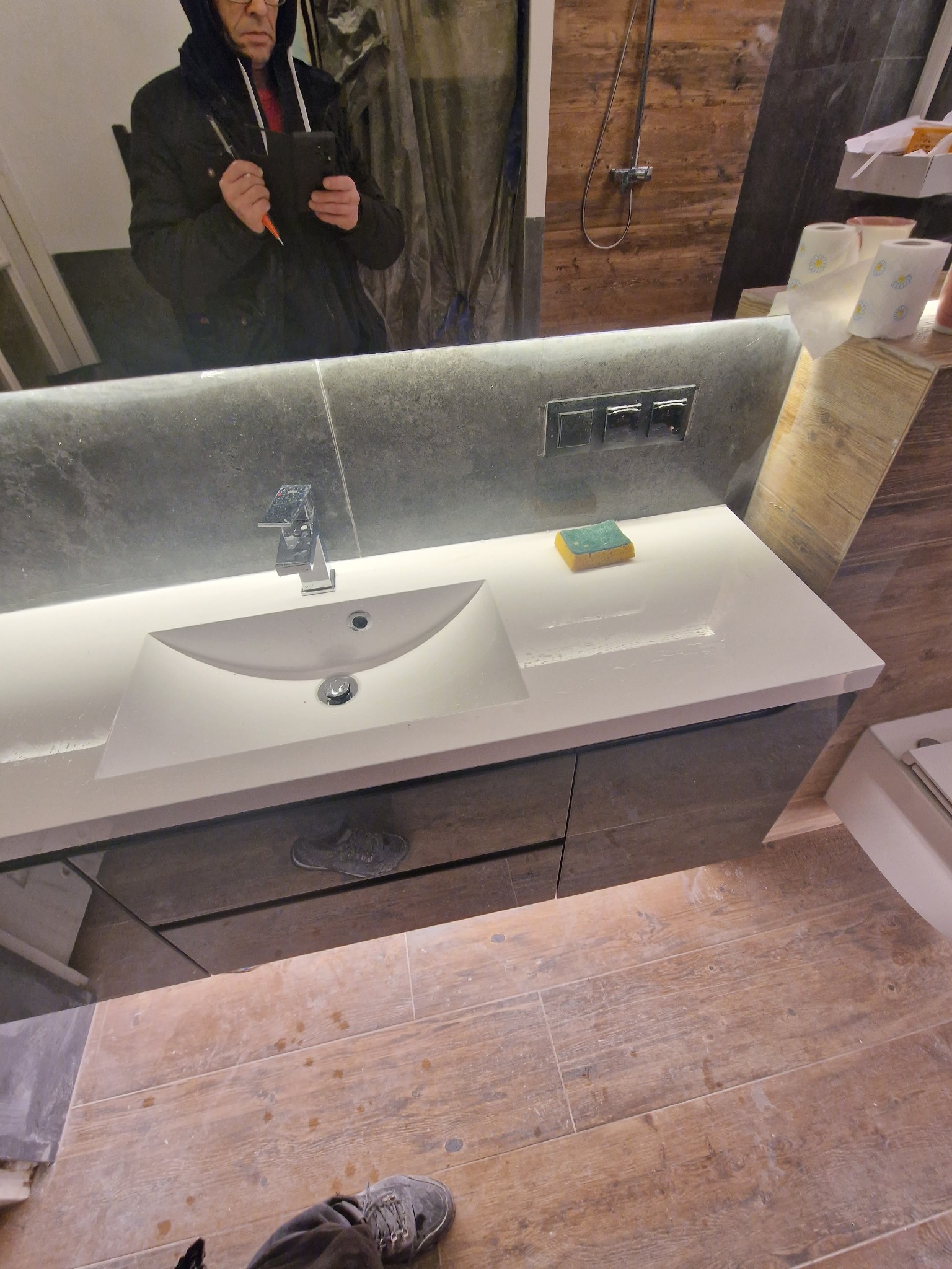 Nowoczesna umywalka z szarą szafką, drewnopodobne płytki podłogowe, odbicie osoby z telefonem w lustrze, widoczne gniazdka elektryczne nad umywalką.