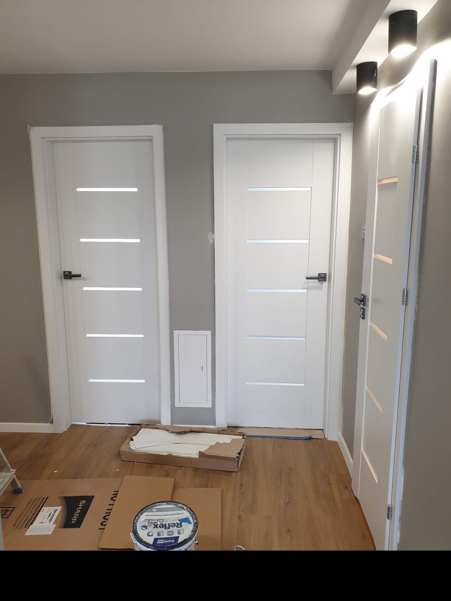 Montaż dwóch białych drzwi wewnętrznych z poziomymi przeszkleniami, widoczna puszka z farbą podkładową Reflex Grunt i kartony ochronne na podłodze.