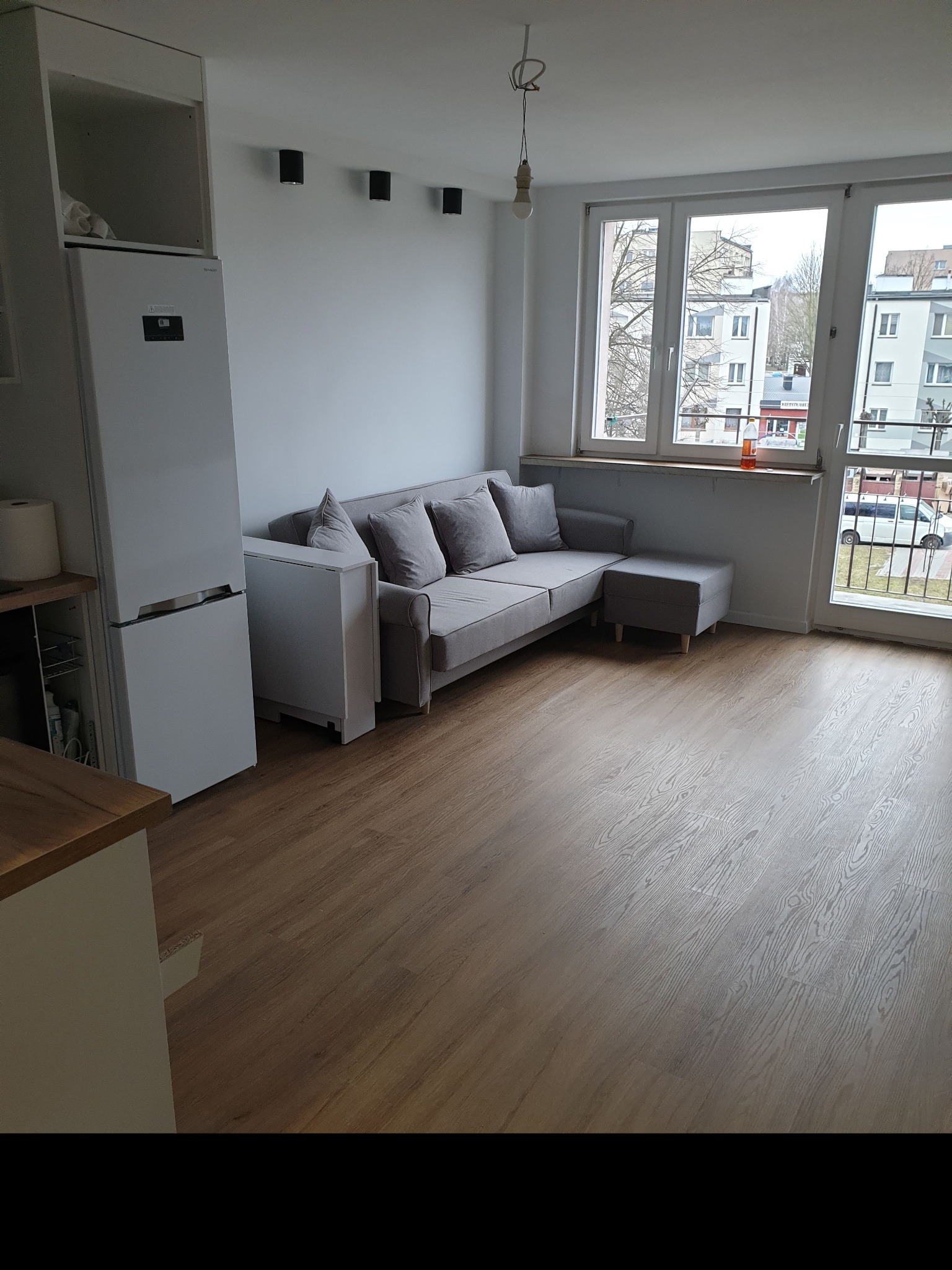 Jasne wnętrze pokoju z szarą sofą z poduszkami, białą lodówką, drewnianą podłogą i dużym oknem balkonowym z widokiem na budynki na zewnątrz.
