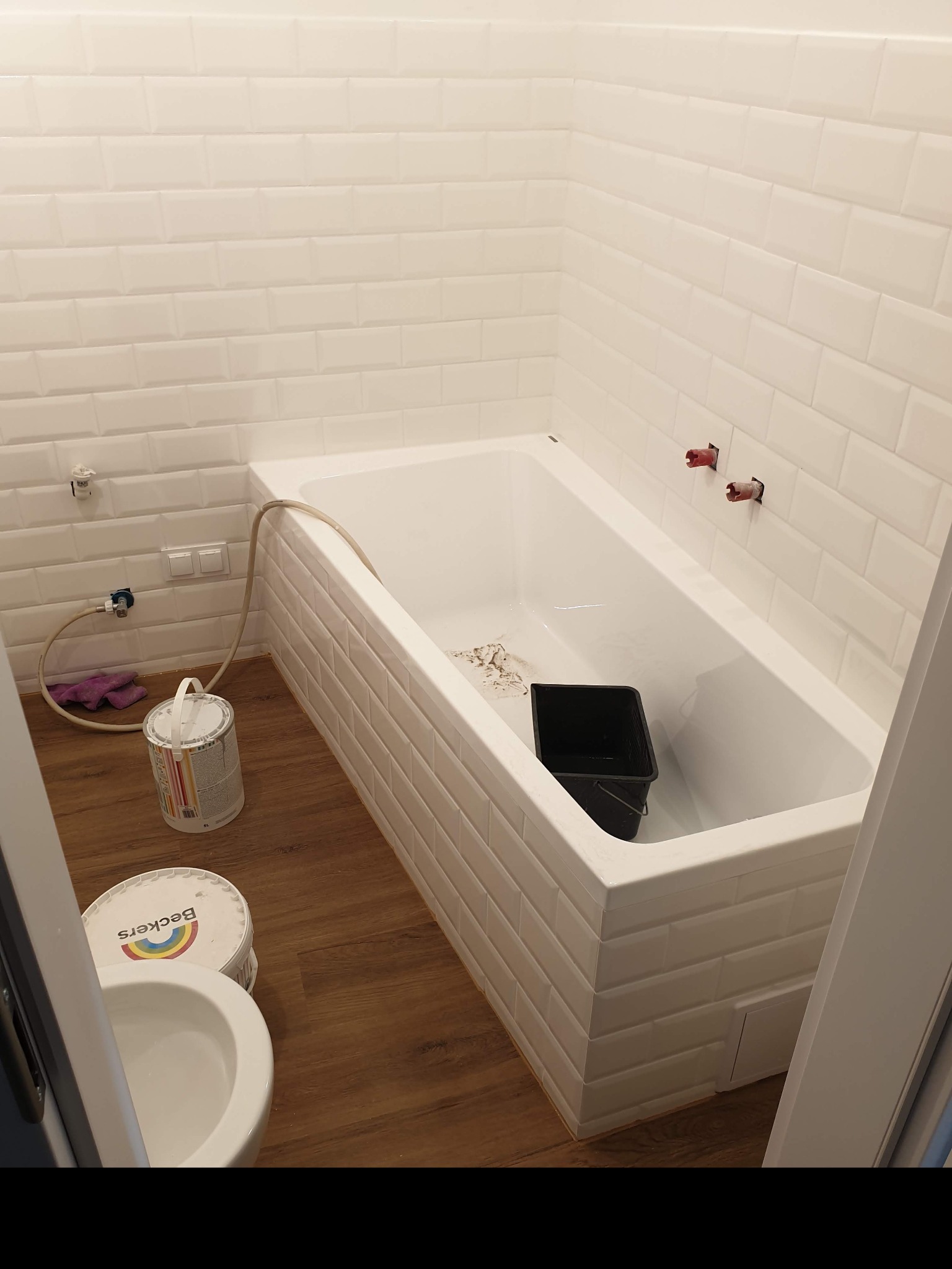 Wnętrze łazienki w trakcie prac: biała wanna obłożona kafelkami typu metro, wiadro wewnątrz, na podłodze puszki farb Beckers oraz fragment toalety.