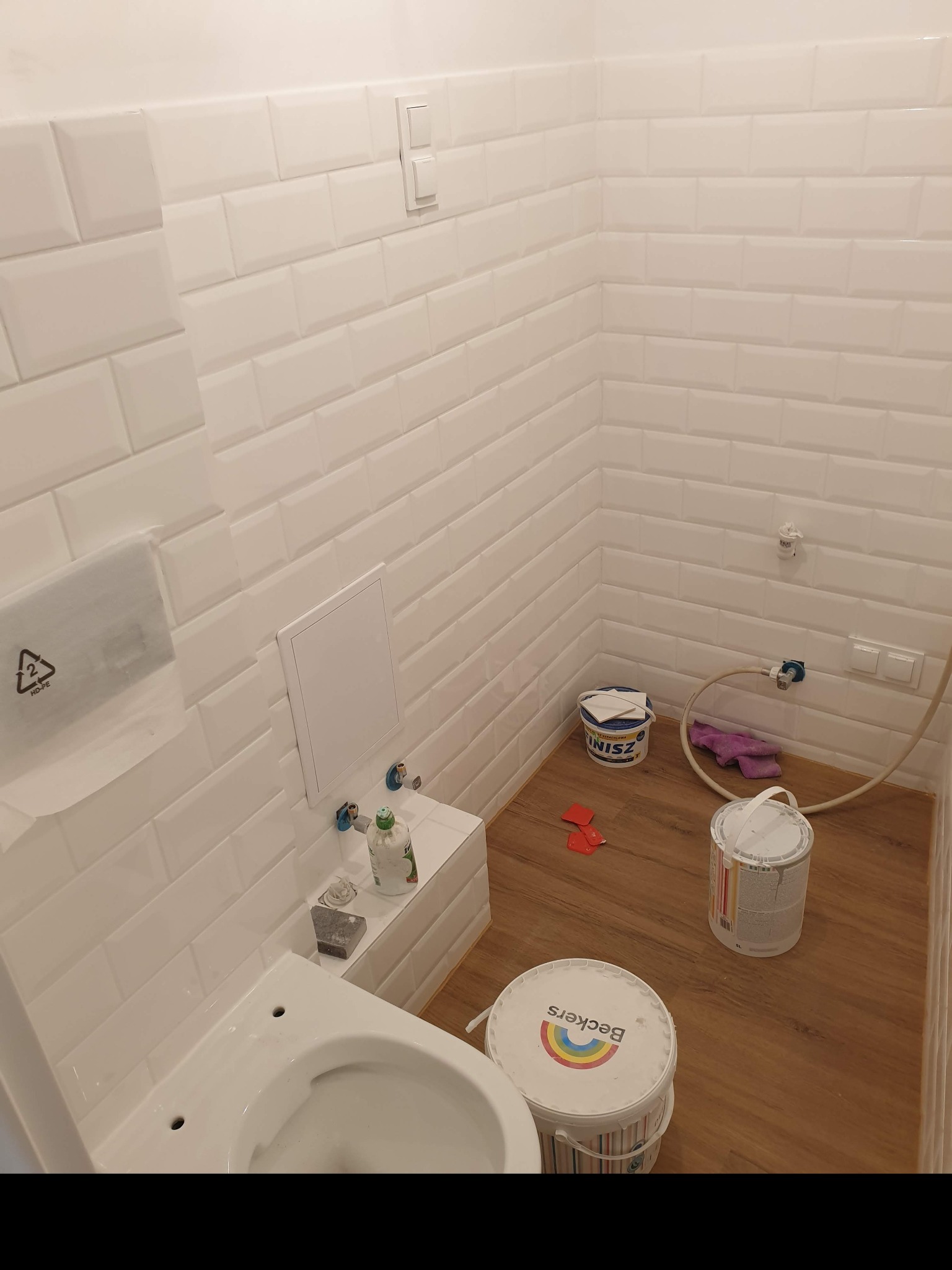 Biała łazienka w trakcie prac wykończeniowych, widoczne białe płytki typu metro, niedokończona toaleta, wiadra z farbą Beckers i Inisz oraz narzędzia glazurnicze na podłodze imitującej drewno.