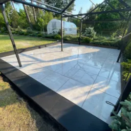 Nowoczesny taras z jasnych, kwadratowych płytek ceramicznych, otoczony czarną obwódką i metalową konstrukcją pergoli, z widokiem na zadbany ogród z iglakami i szklarnią w tle.