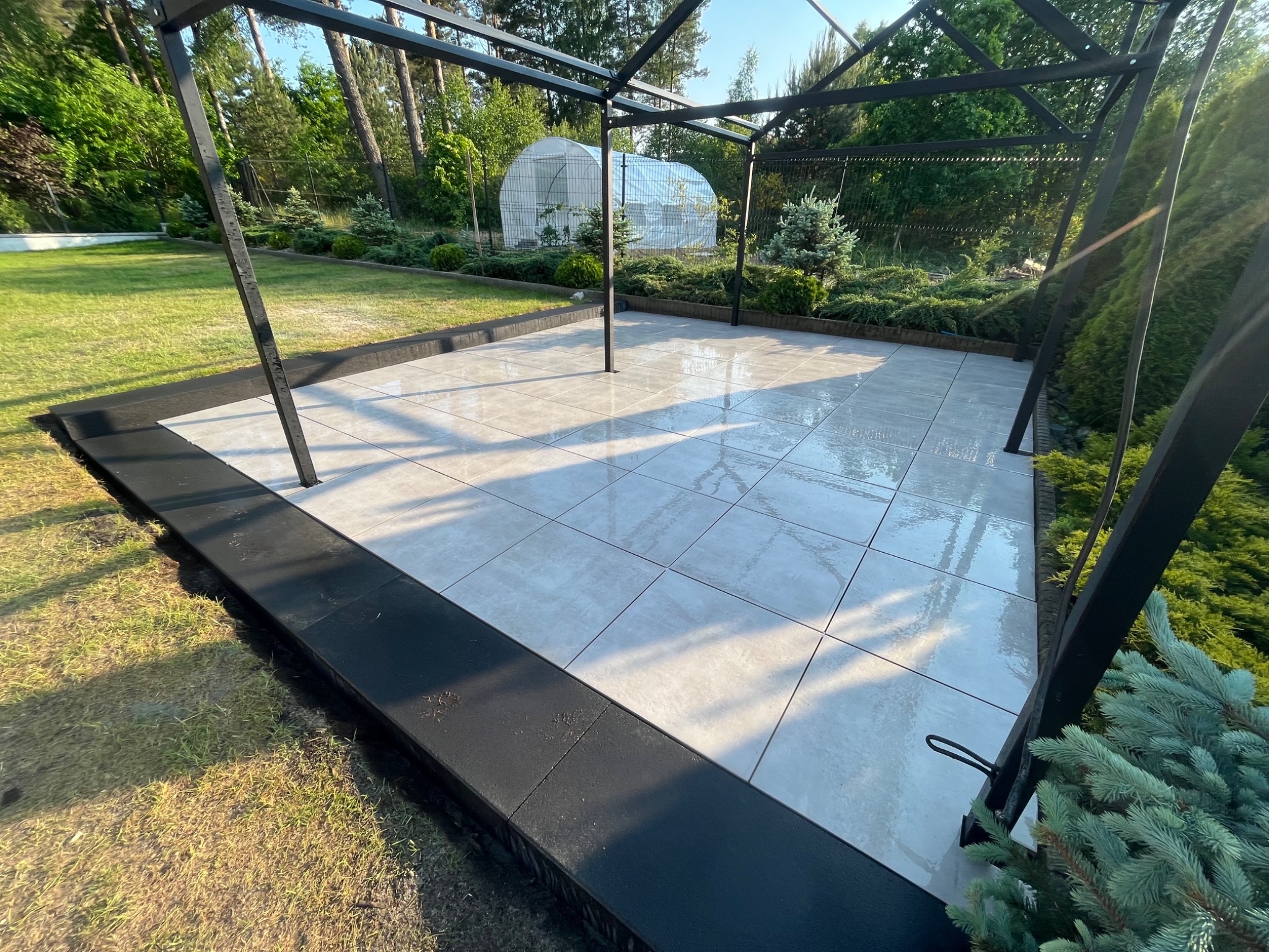 Nowoczesny taras z jasnych, kwadratowych płytek ceramicznych, otoczony czarną obwódką i metalową konstrukcją pergoli, z widokiem na zadbany ogród z iglakami i szklarnią w tle.