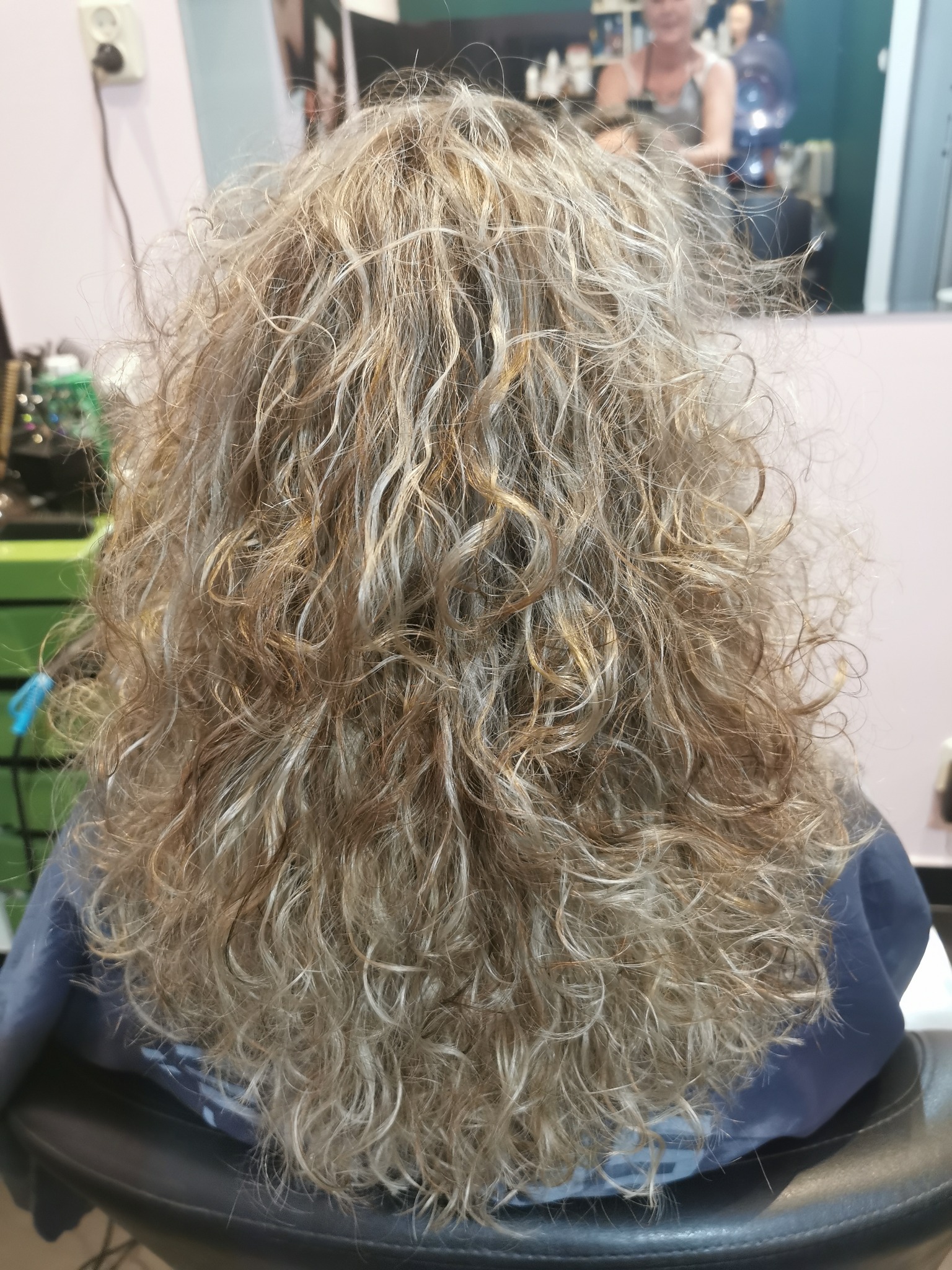 Tył głowy z kręconymi, blond włosami z odrostami, widziany w salonie, klientka siedzi na czarnym fotelu fryzjerskim, okryta peleryną.