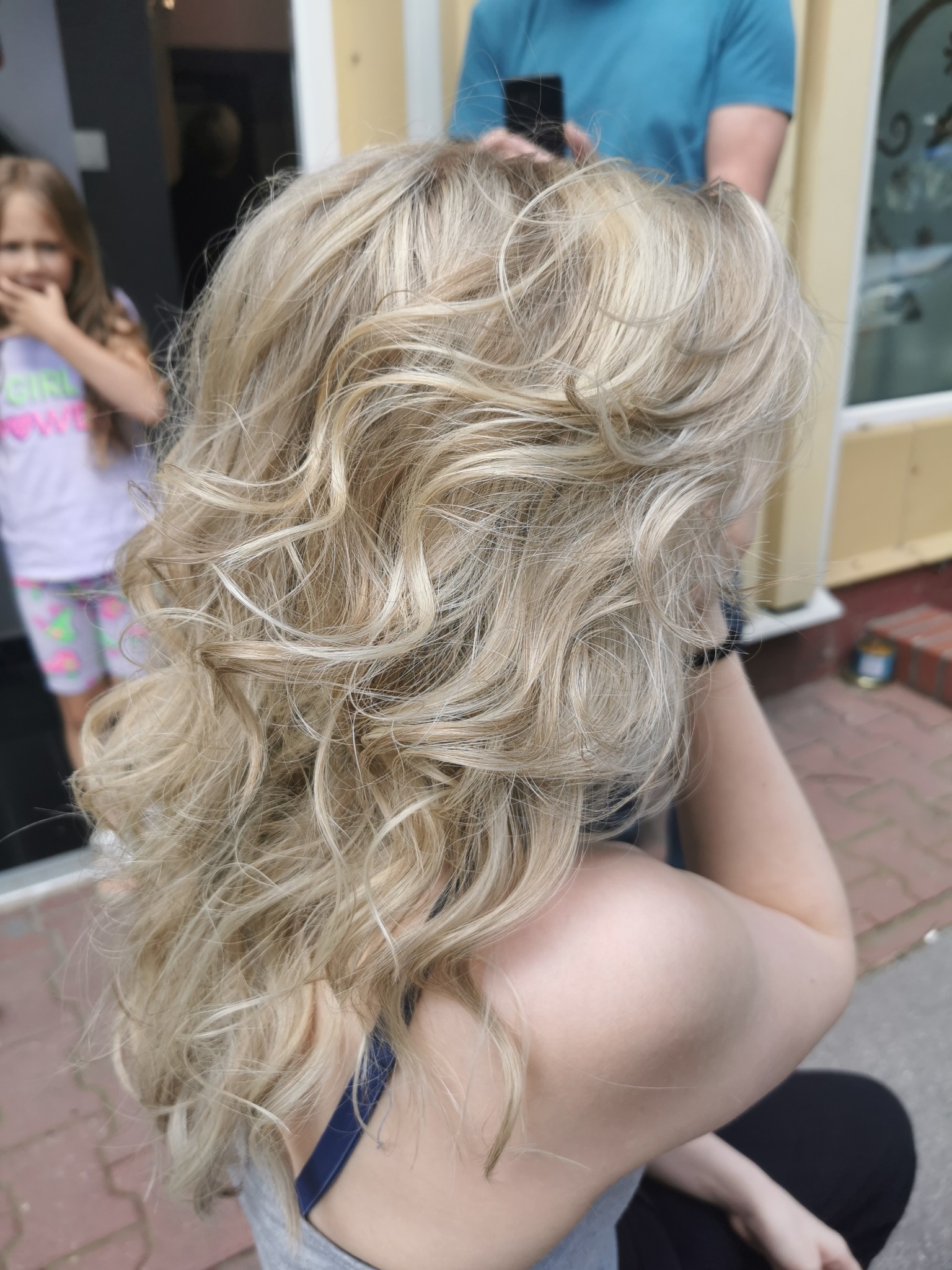 Blond włosy z delikatnymi lokami, widoczne zza ramienia siedzącej kobiety, w tle fragment budynku i osoba z telefonem.