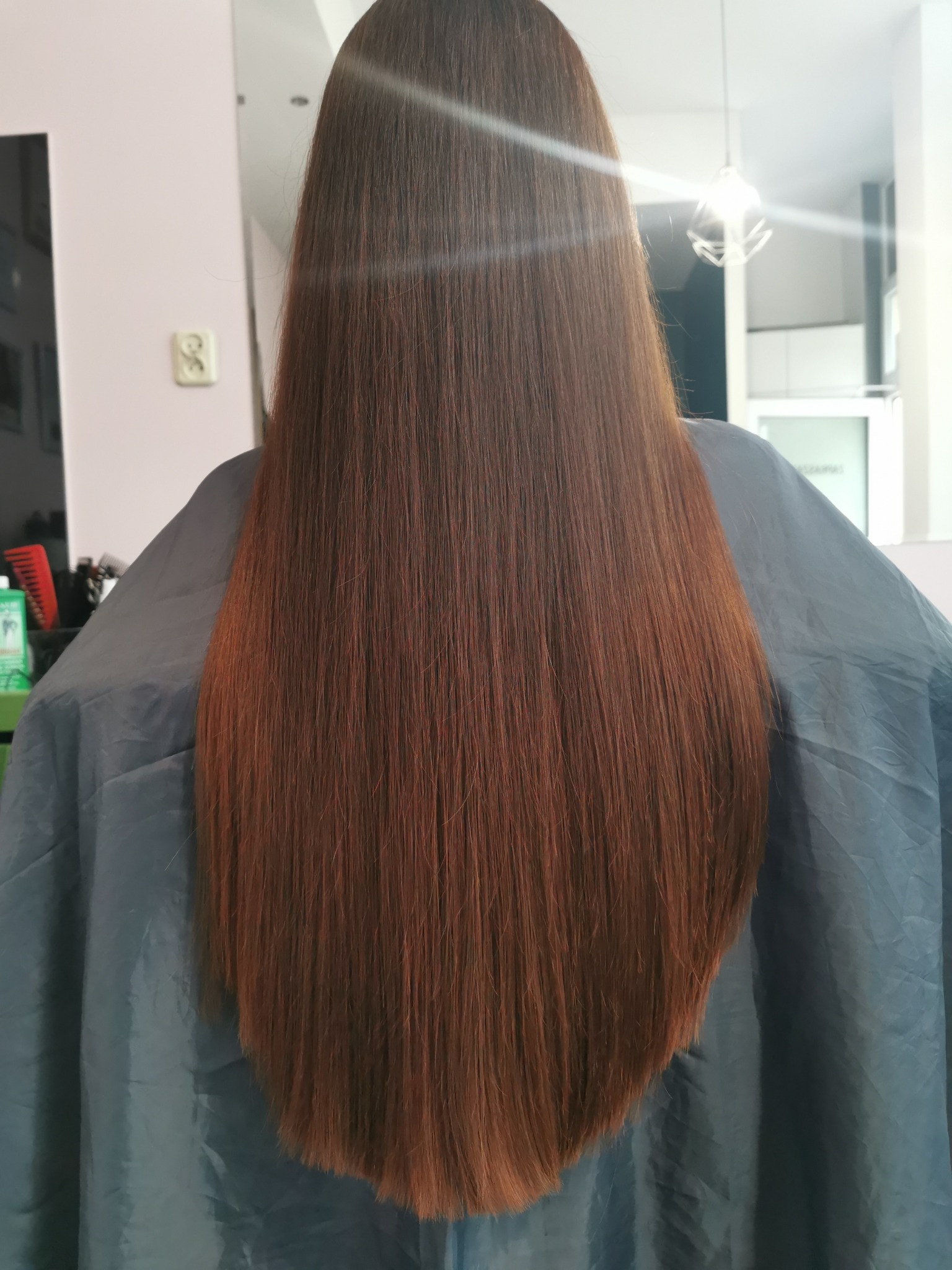 Długie, proste, kasztanowe włosy klientki widziane od tyłu, osłonięte peleryną w salonie.
