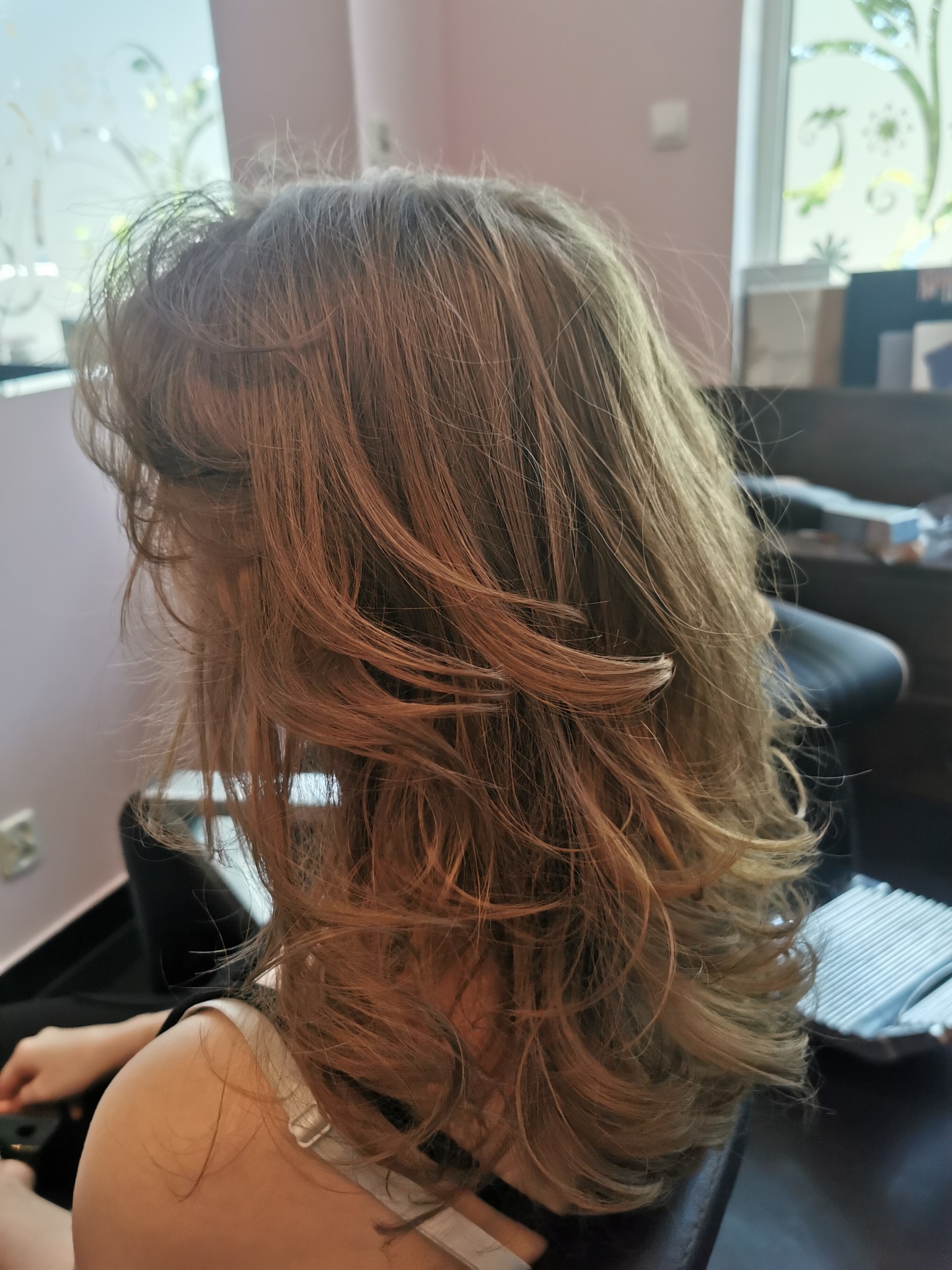 Uczesana, pofalowana fryzura w odcieniach blondu i brązu, widok z boku na fotelu w salonie, z delikatnymi refleksami i układającymi się warstwami.