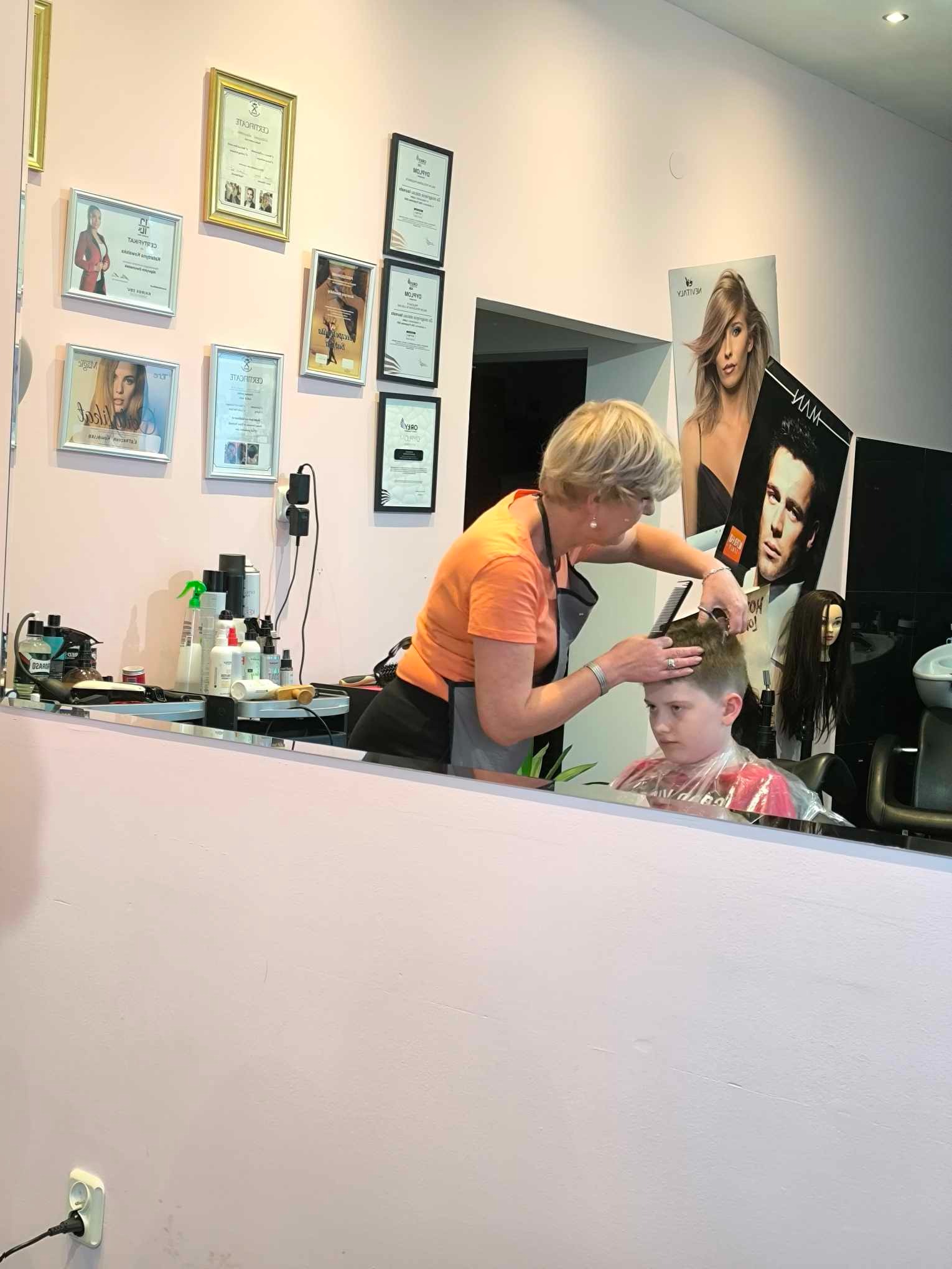 Kobieta w pomarańczowej koszulce, z krótkimi blond włosami, strzyże młodego chłopca w salonie, na ścianie wiszą oprawione certyfikaty i plakaty reklamowe.