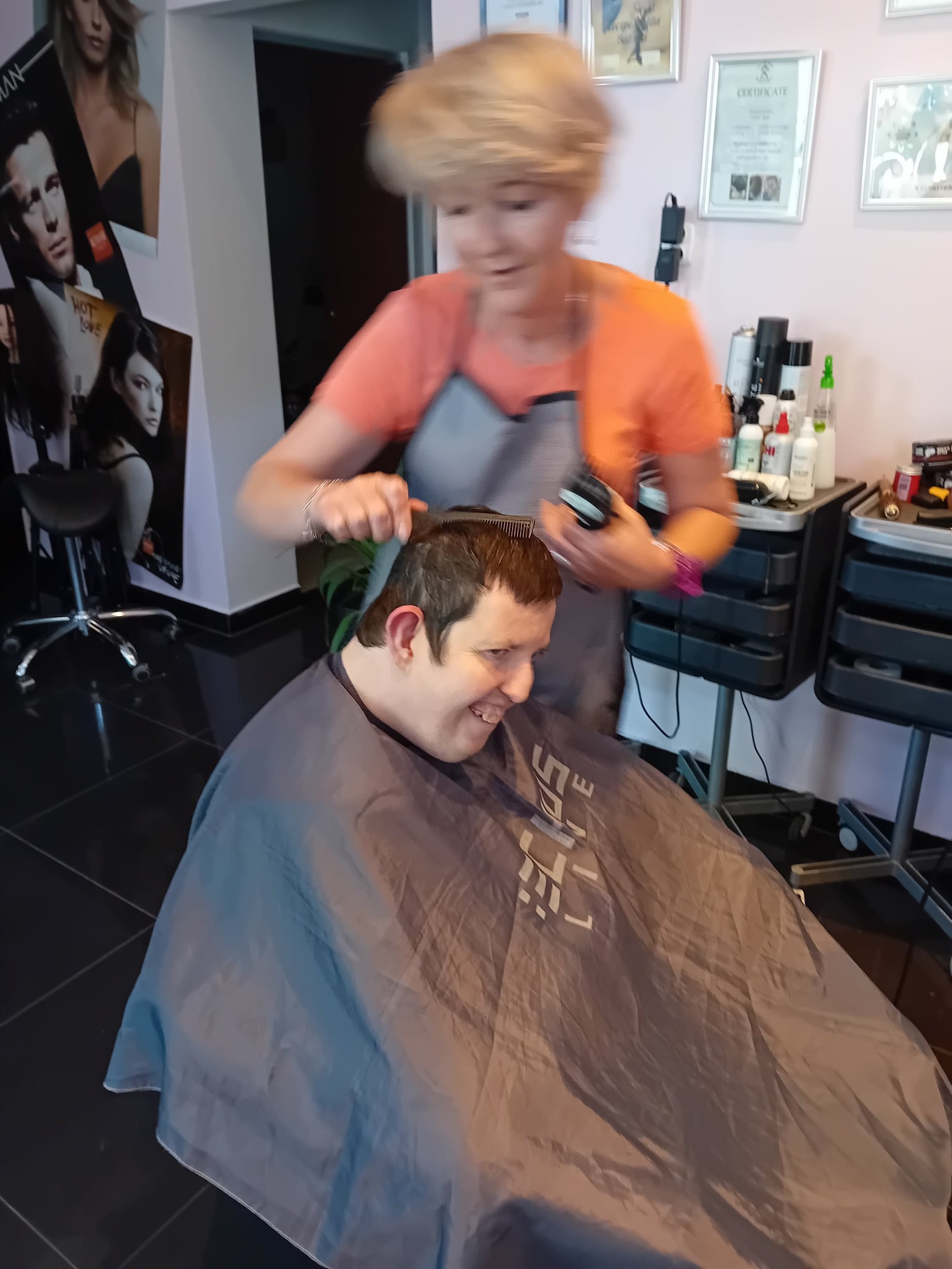 Klient uśmiecha się podczas strzyżenia włosów w salonie, fryzjerka w pomarańczowej koszulce i szarym fartuchu używa maszynki i grzebienia, w tle plakaty i kosmetyki.