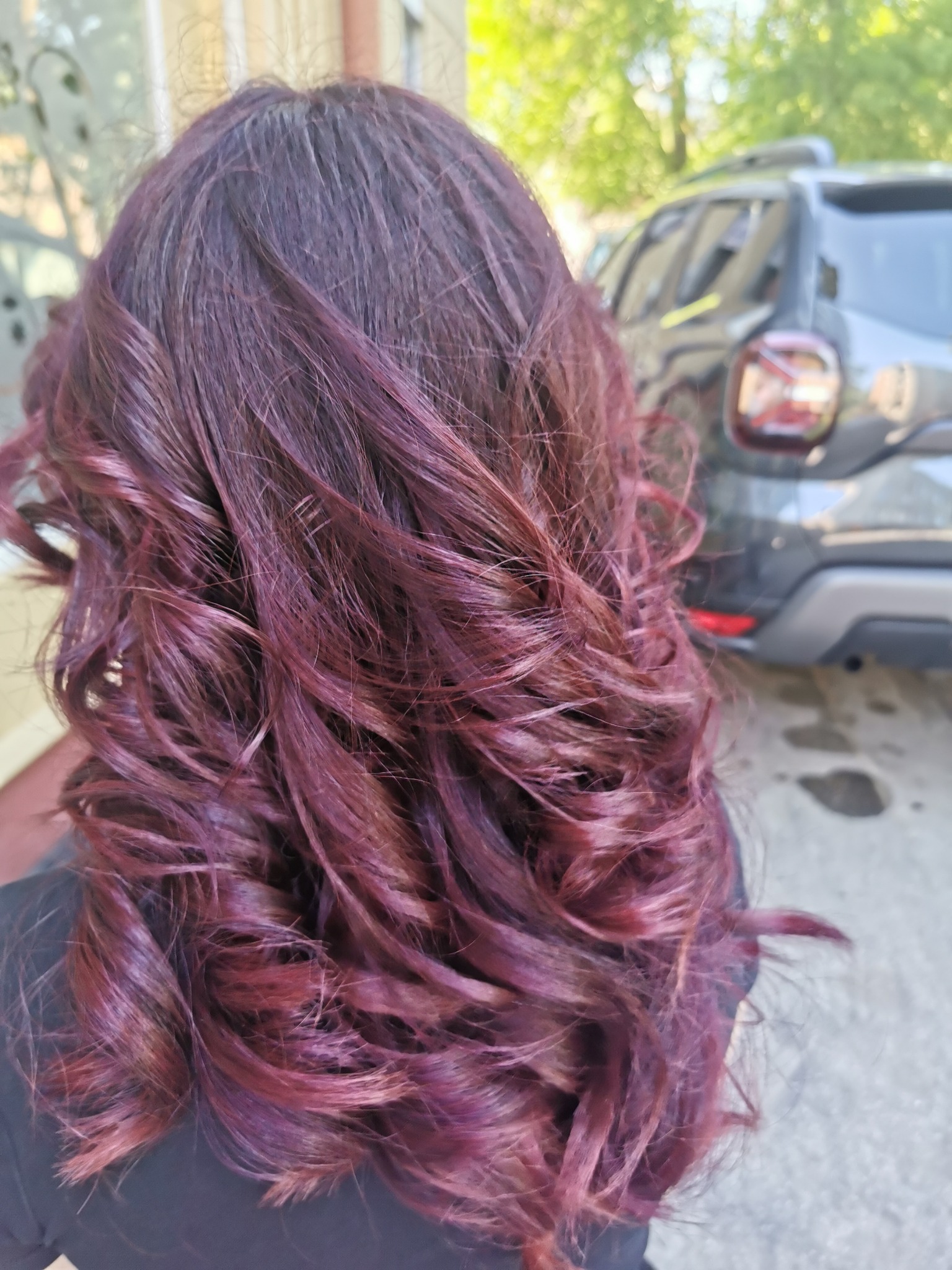Tył głowy z falowanymi włosami w odcieniach fioletu i bordo, z widocznymi refleksami, na rozmytym tle samochodu i zieleni.