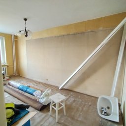 Vitalii Mordvinov - Pokój w trakcie przygotowania do remontu: zwinięte dywany, białe listwy przypodłogowe oparte o ścianę, mały stołek i zdemontowana umywalka z baterią.