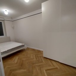 Vitalii Mordvinov - Pusty pokój po remoncie: parkiet w jodełkę, białe ściany z czarnym paskiem pod sufitem, biała szafa w zabudowie, łóżko z materacem owiniętym folią ochronną, dwa punkty oświetleniowe na suficie.