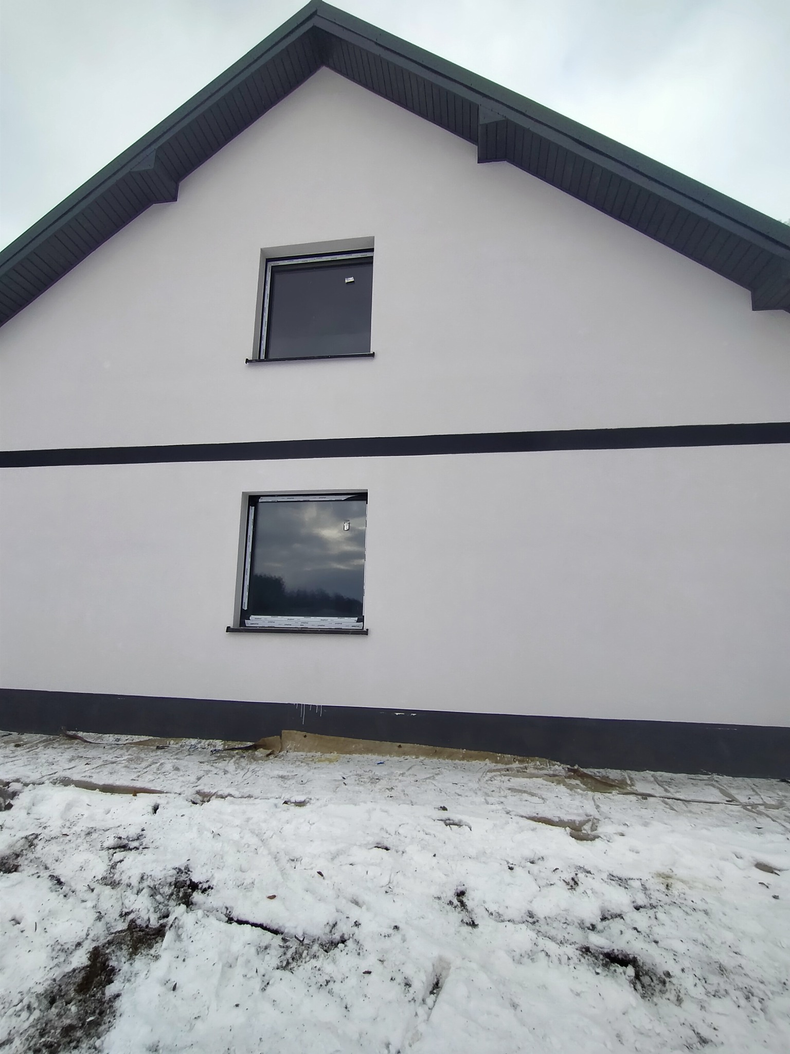Elewacja domu jednorodzinnego po remoncie z dwoma oknami w czarnych ramach, świeżo pomalowana na biało z ciemnoszarymi akcentami na dachu i cokole, widoczne ślady śniegu na ziemi.