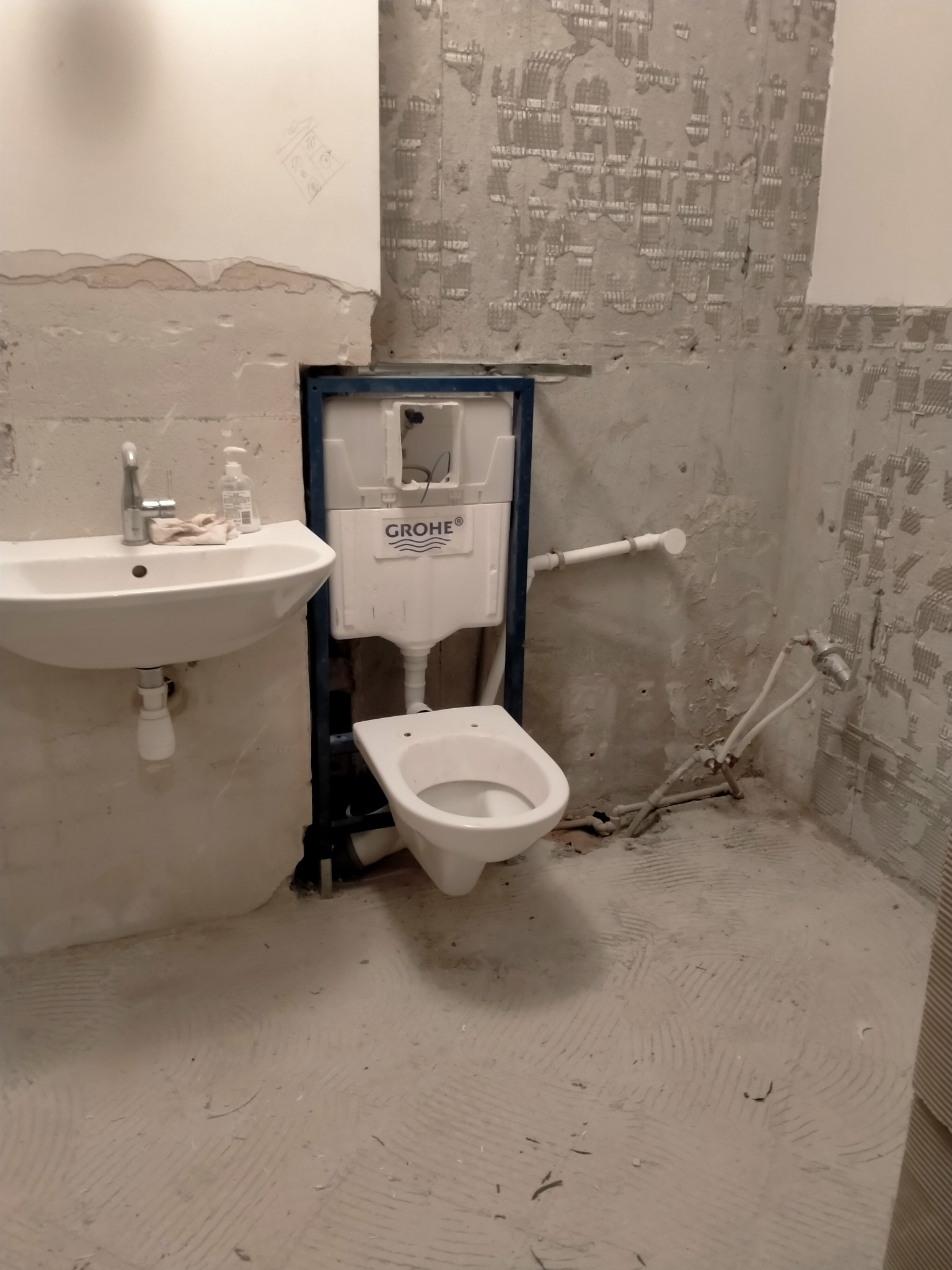Łazienka w trakcie remontu: zamontowana toaleta Grohe na stelażu, umywalka z baterią, ściany przygotowane pod płytki, widoczne instalacje wodne.