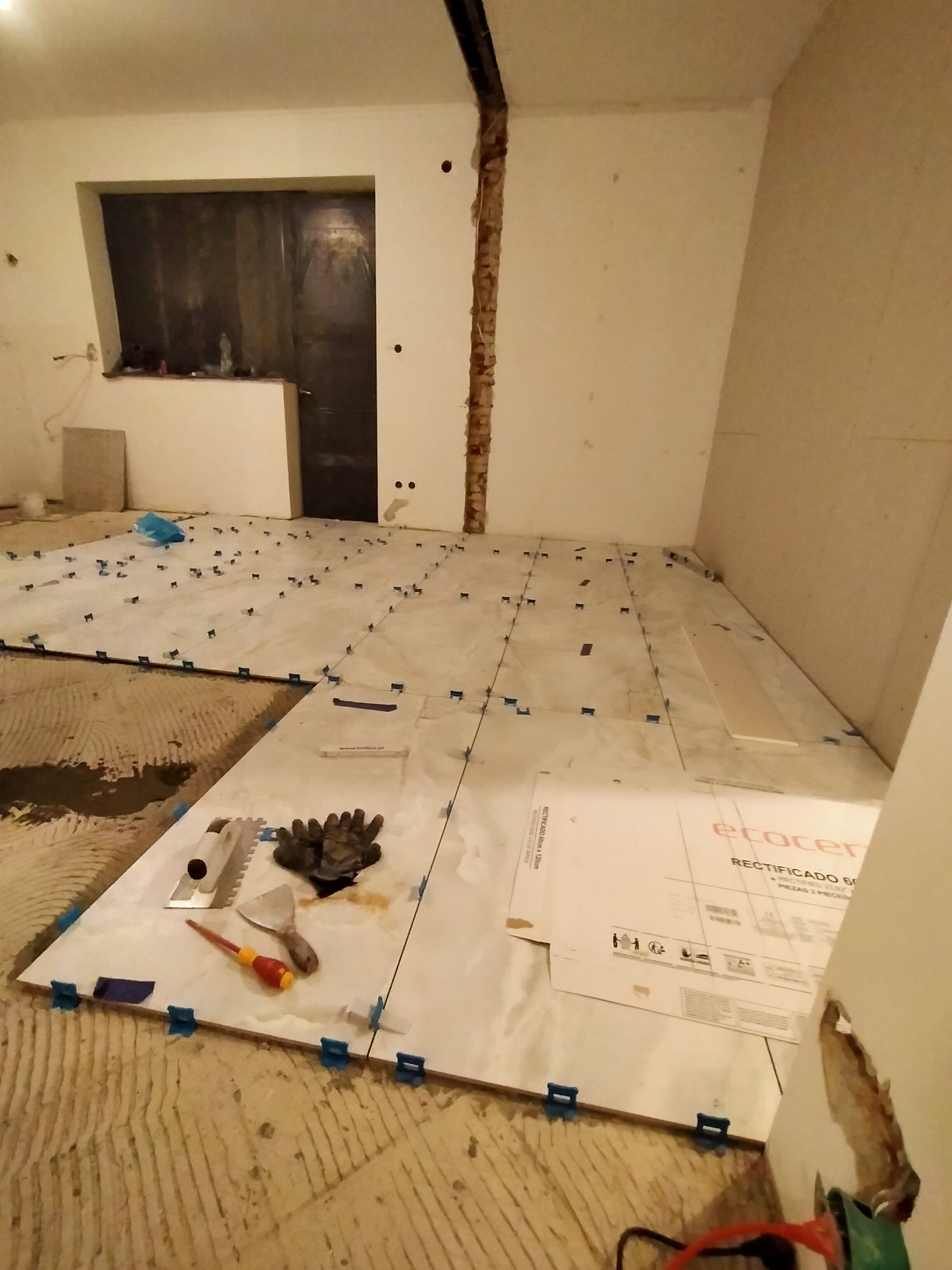 Wnętrze w trakcie układania jasnych, kwadratowych płytek podłogowych z użyciem niebieskich klipsów poziomujących; widoczne narzędzia glazurnicze, rękawice robocze i karton z nazwą producenta płytek.