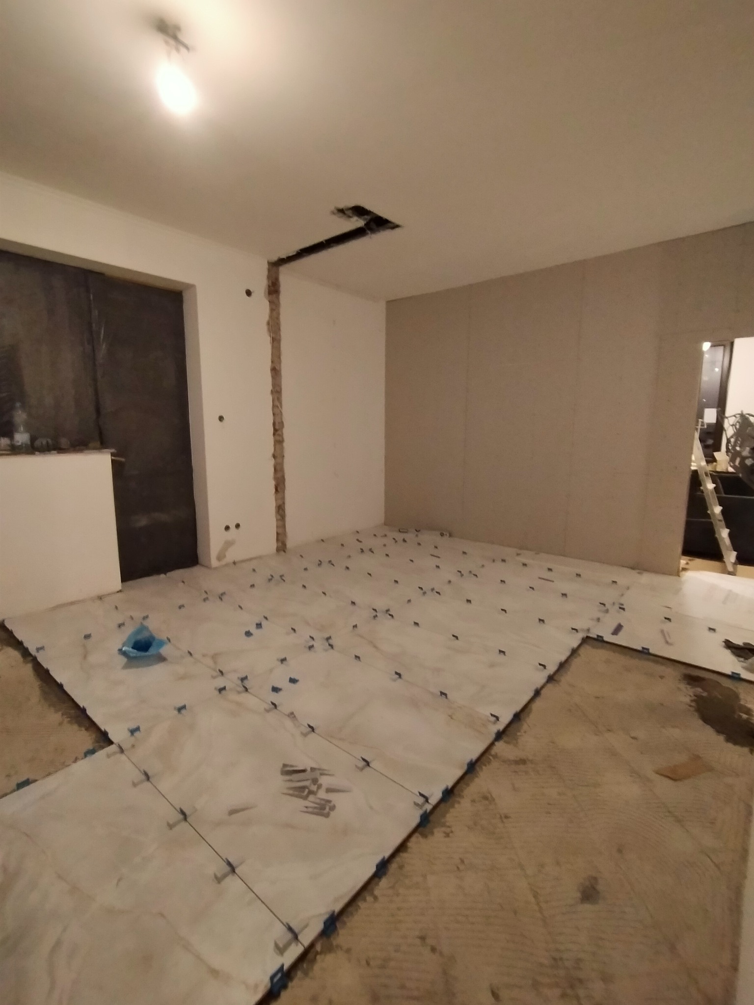 Wnętrze w trakcie prac z układaniem płytek podłogowych, widoczne płytki z krzyżykami dystansowymi, przygotowana ściana z płyt kartonowo-gipsowych i odkryta instalacja elektryczna w suficie.