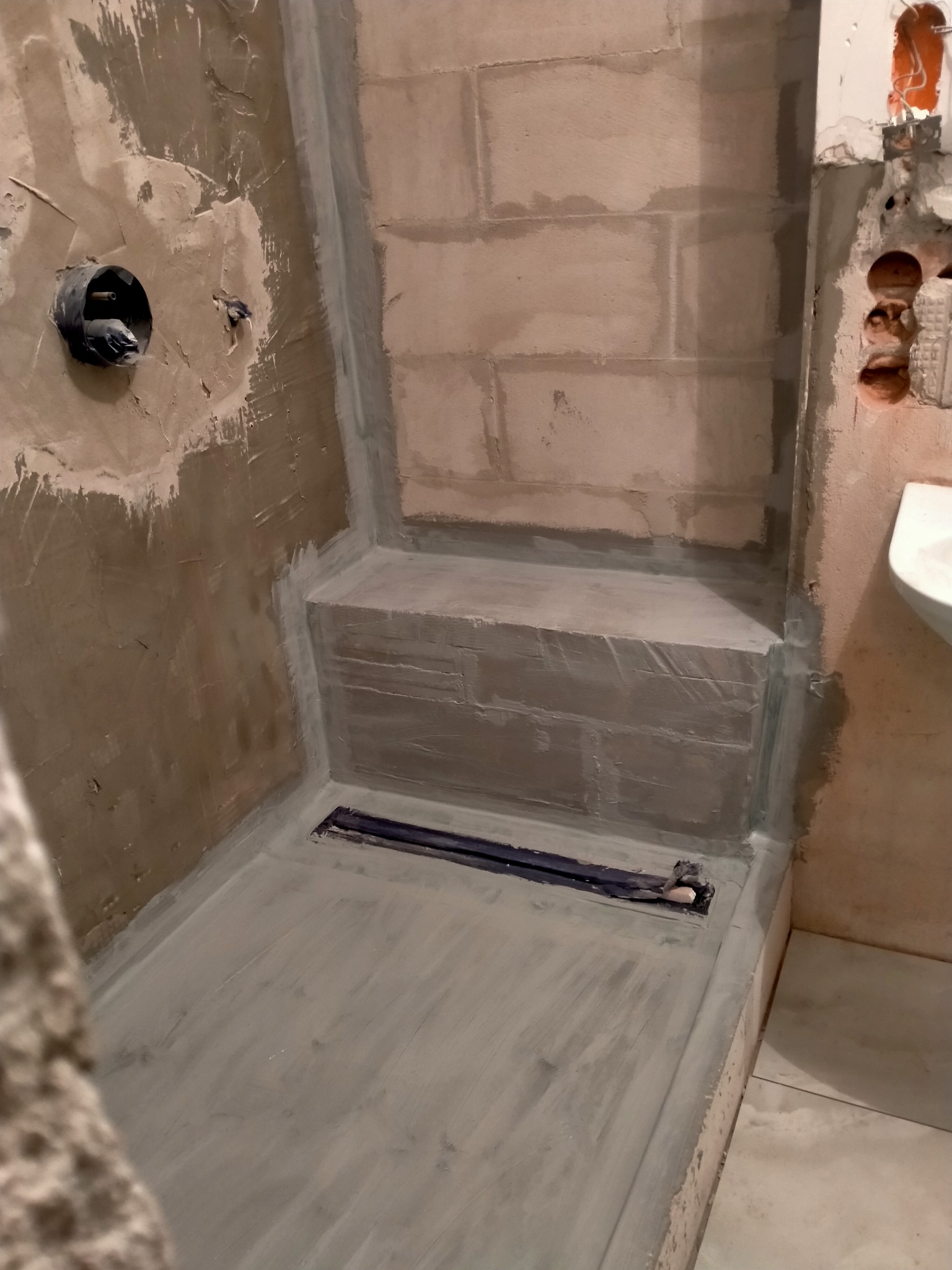 Przygotowanie do hydroizolacji w kabinie prysznicowej: surowe ściany z betonu komórkowego, zabezpieczone narożniki i odpływ liniowy w posadzce, widoczna instalacja elektryczna.