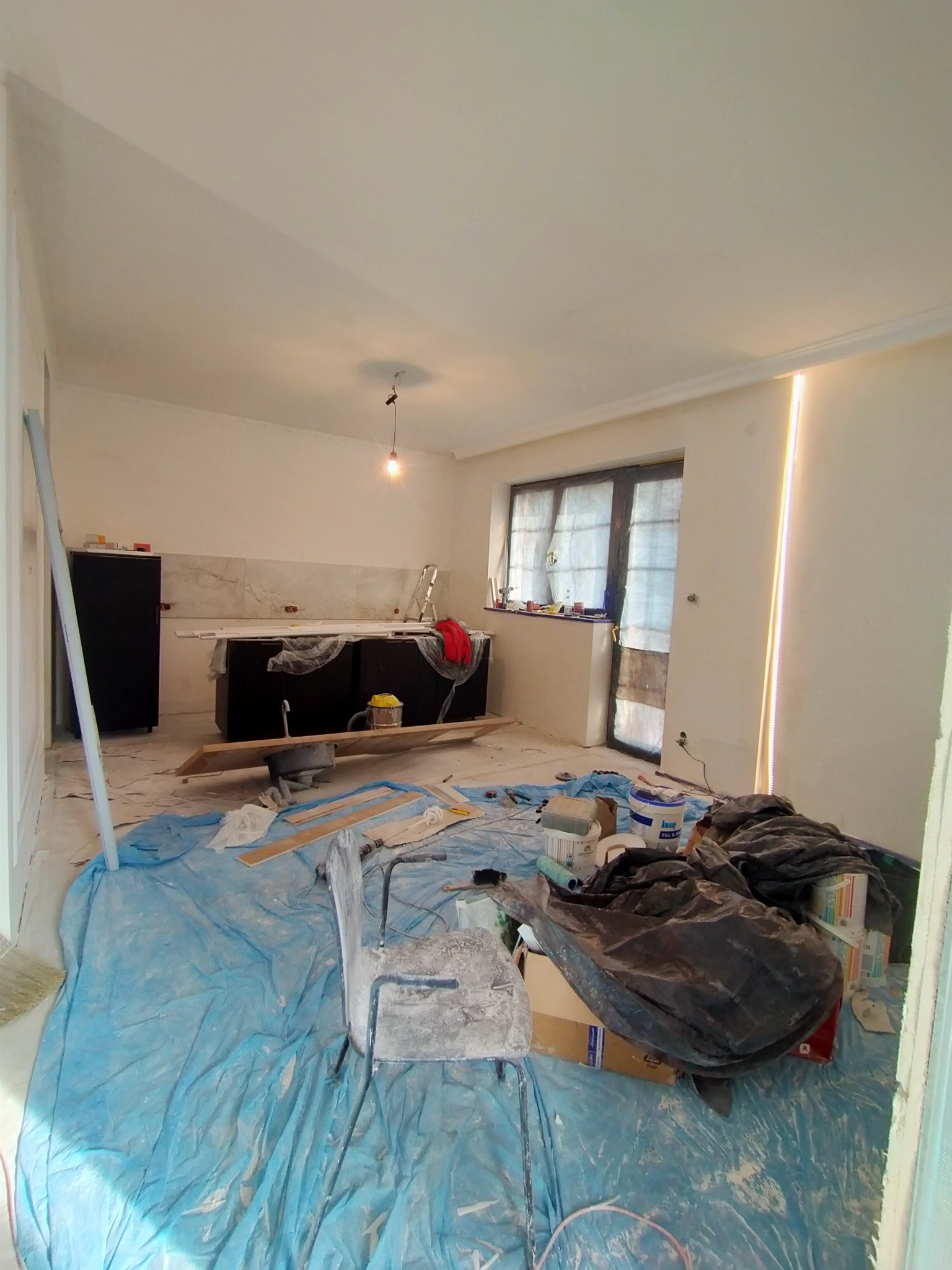 Wnętrze kuchni w trakcie prac wykończeniowych: zabezpieczona folią podłoga, widoczne narzędzia, pędzle, wiadro z farbą, niedokończone ściany i położone płytki nad blatem kuchennym.