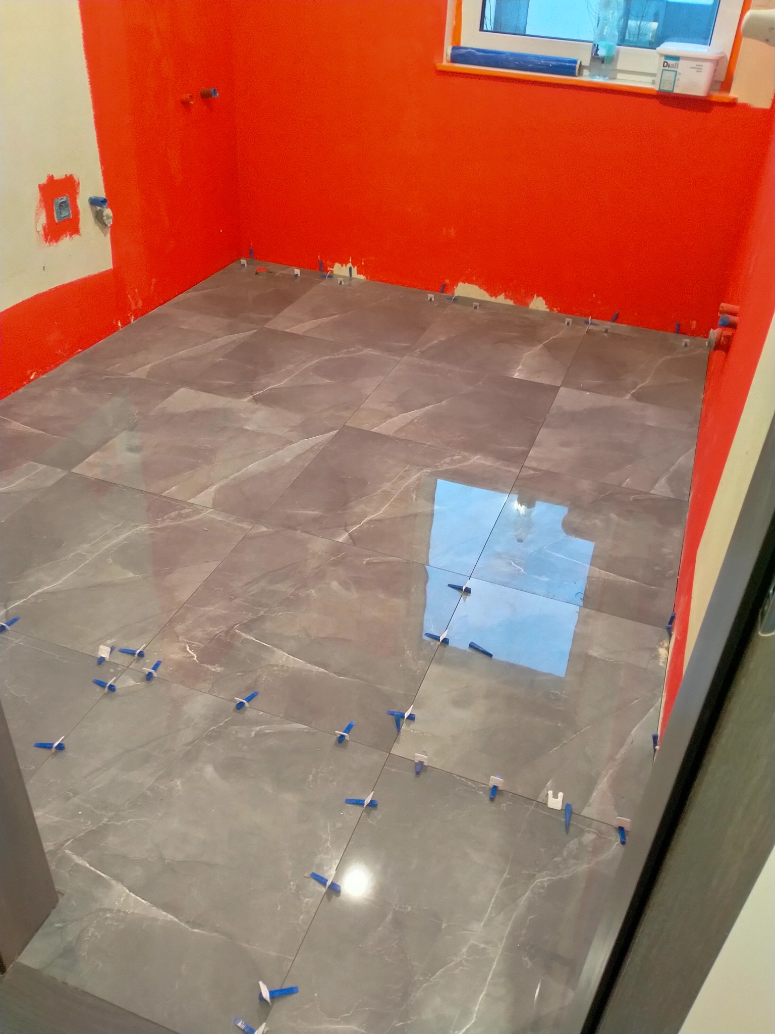 Szare płytki podłogowe imitujące marmur, ułożone w trakcie prac, z użyciem niebieskich i białych krzyżyków dystansowych, ściany pomalowane na jaskrawy pomarańczowy kolor, widoczne instalacje...
