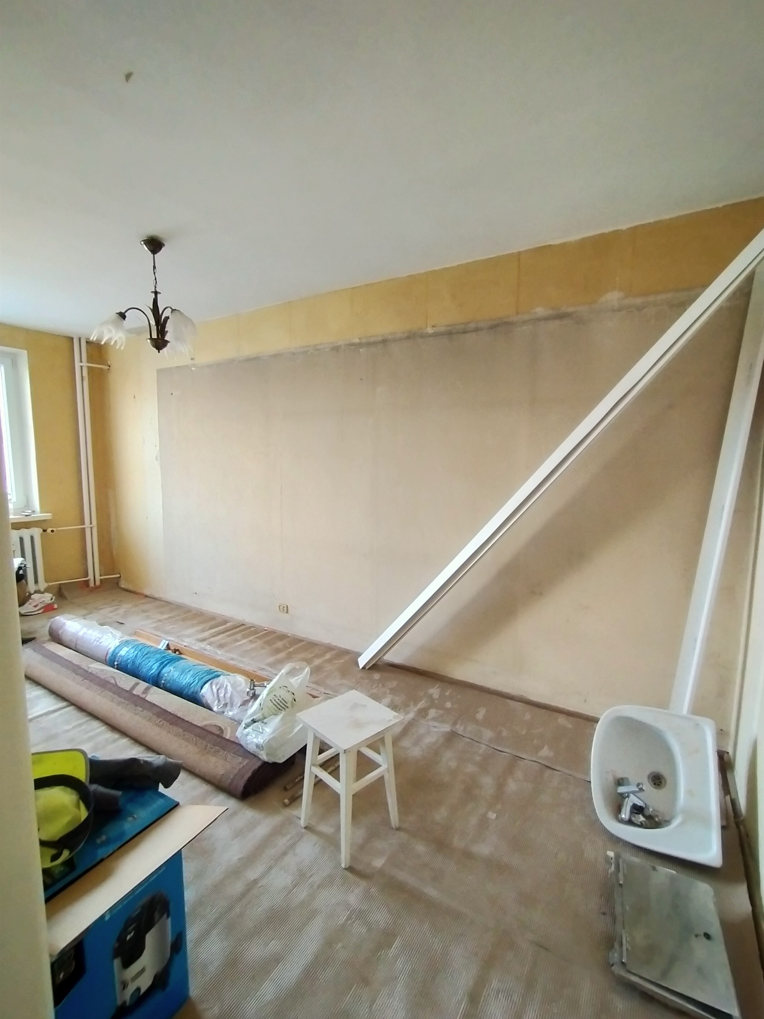 Pokój w trakcie przygotowania do remontu: zwinięte dywany, białe listwy przypodłogowe oparte o ścianę, mały stołek i zdemontowana umywalka z baterią.