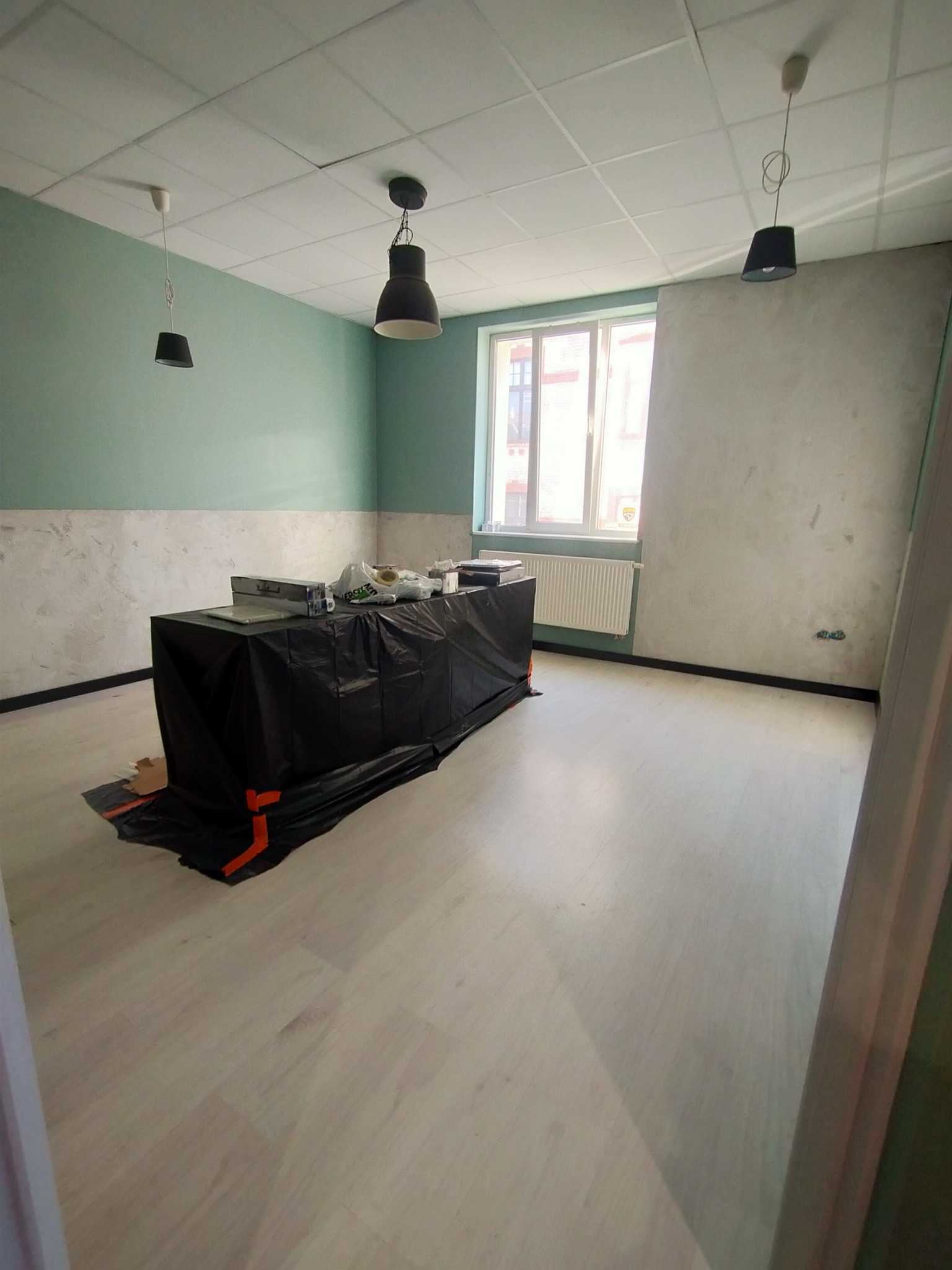 Pokój w trakcie malowania z zabezpieczonymi meblami czarną folią, świeżo pomalowana zielona ściana w górnej części i szara w dolnej. Widoczne trzy lampy sufitowe i okno z grzejnikiem.