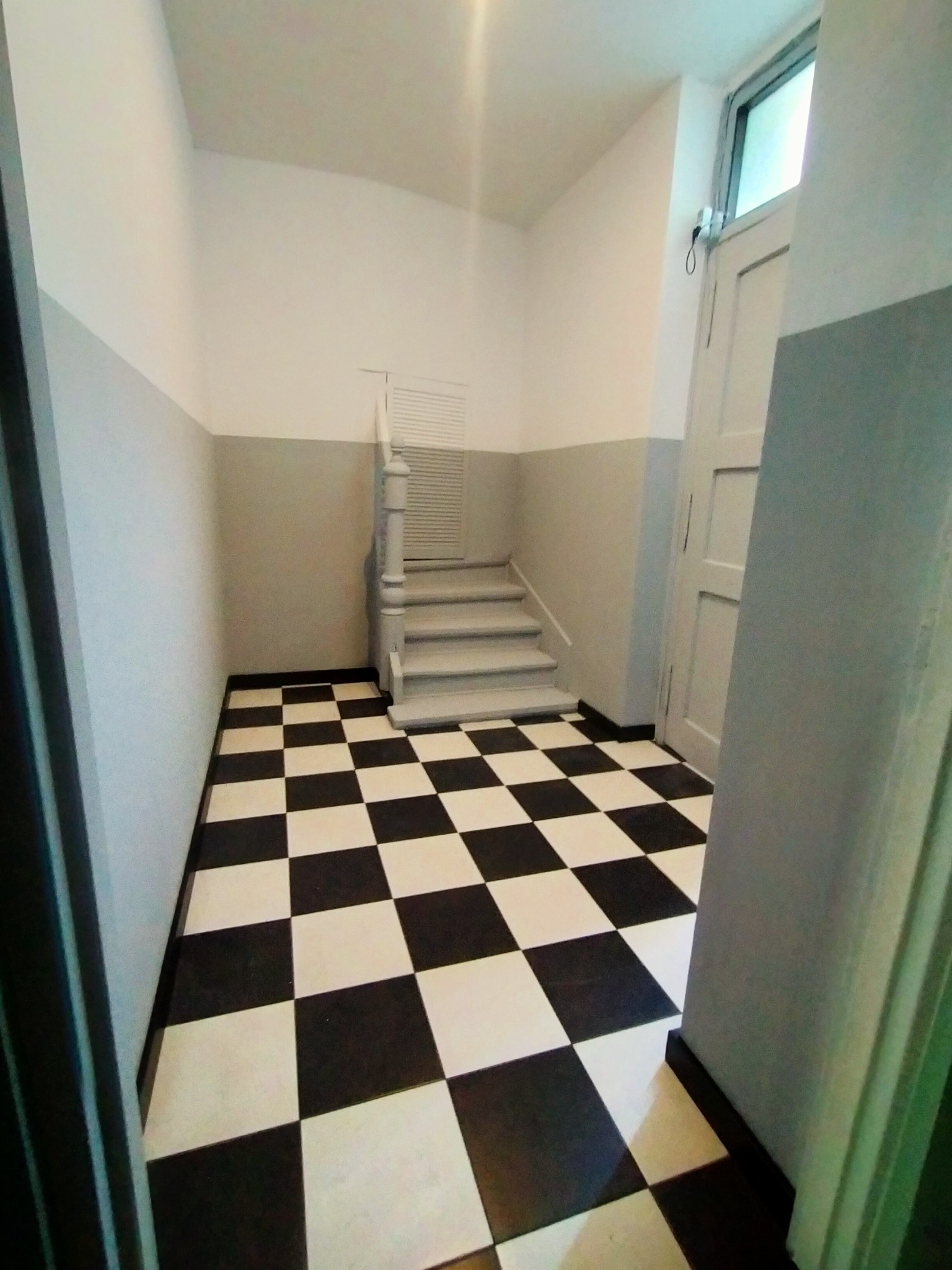 Korytarz z czarno-białą szachownicą na podłodze, ściany pomalowane na biało i szaro, widoczne schody z białą balustradą i białe drzwi.
