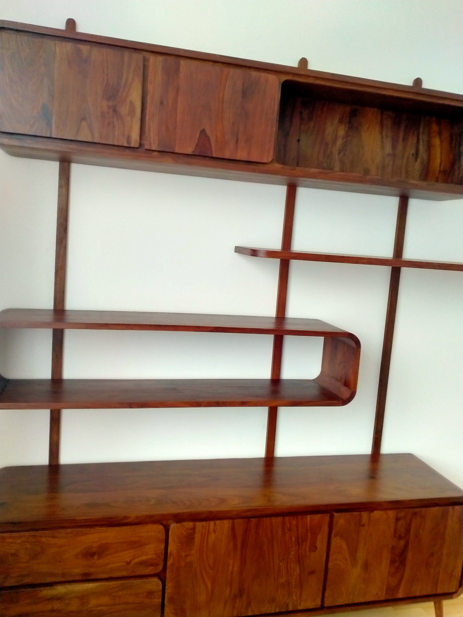 Ręcznie robiony regał z ciemnego drewna z zaokrąglonymi półkami i szafkami na tle białej ściany.