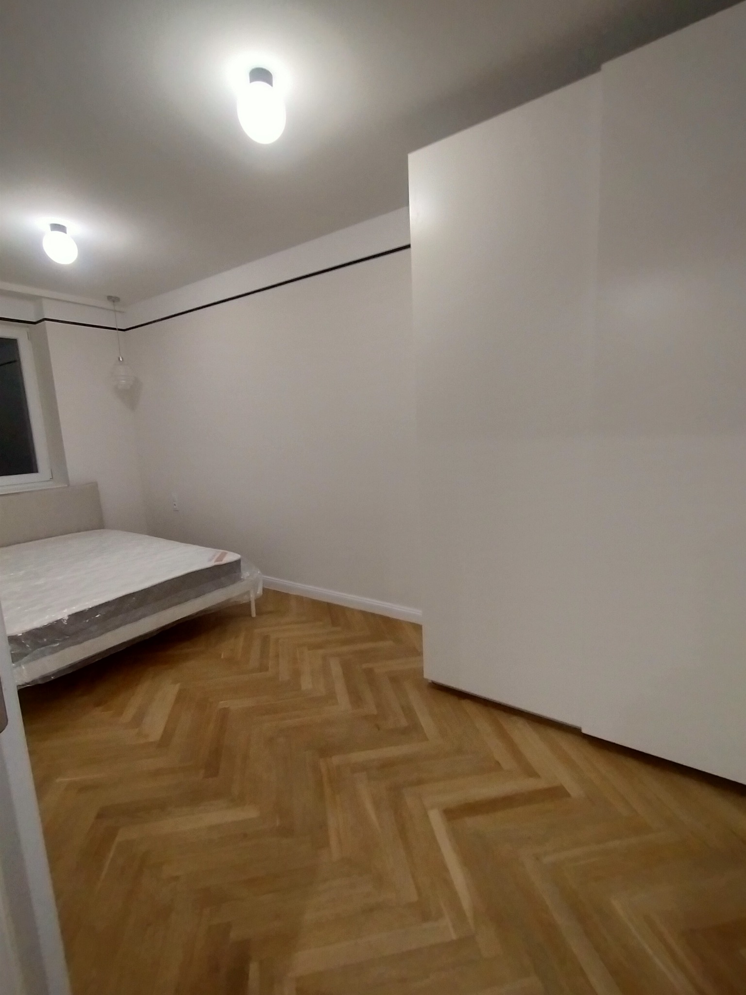 Pusty pokój po remoncie: parkiet w jodełkę, białe ściany z czarnym paskiem pod sufitem, biała szafa w zabudowie, łóżko z materacem owiniętym folią ochronną, dwa punkty oświetleniowe na suficie.
