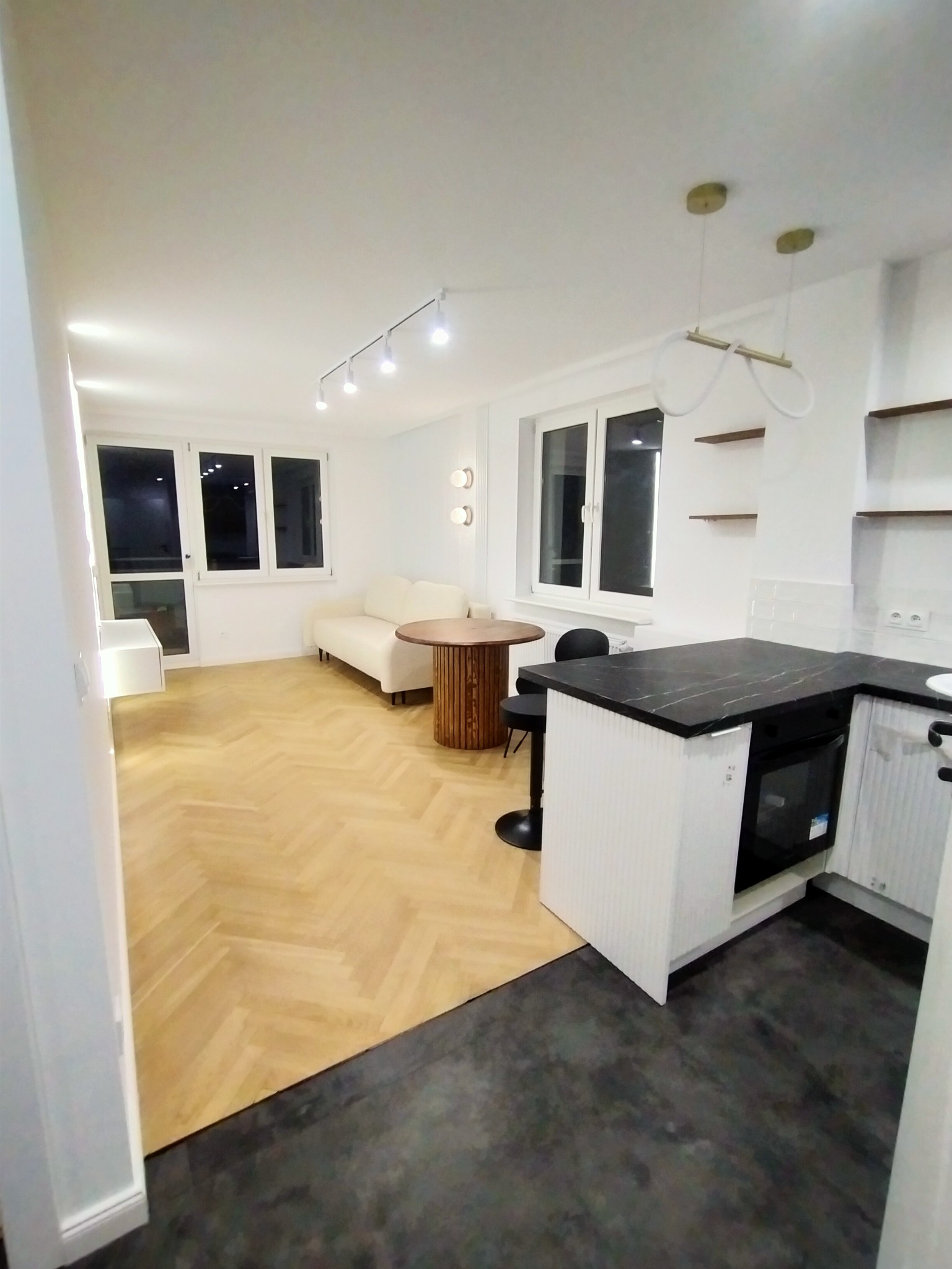 Jasne wnętrze mieszkania po remoncie: parkiet ułożony w jodełkę, białe ściany, minimalistyczne meble i czarny blat kuchenny.