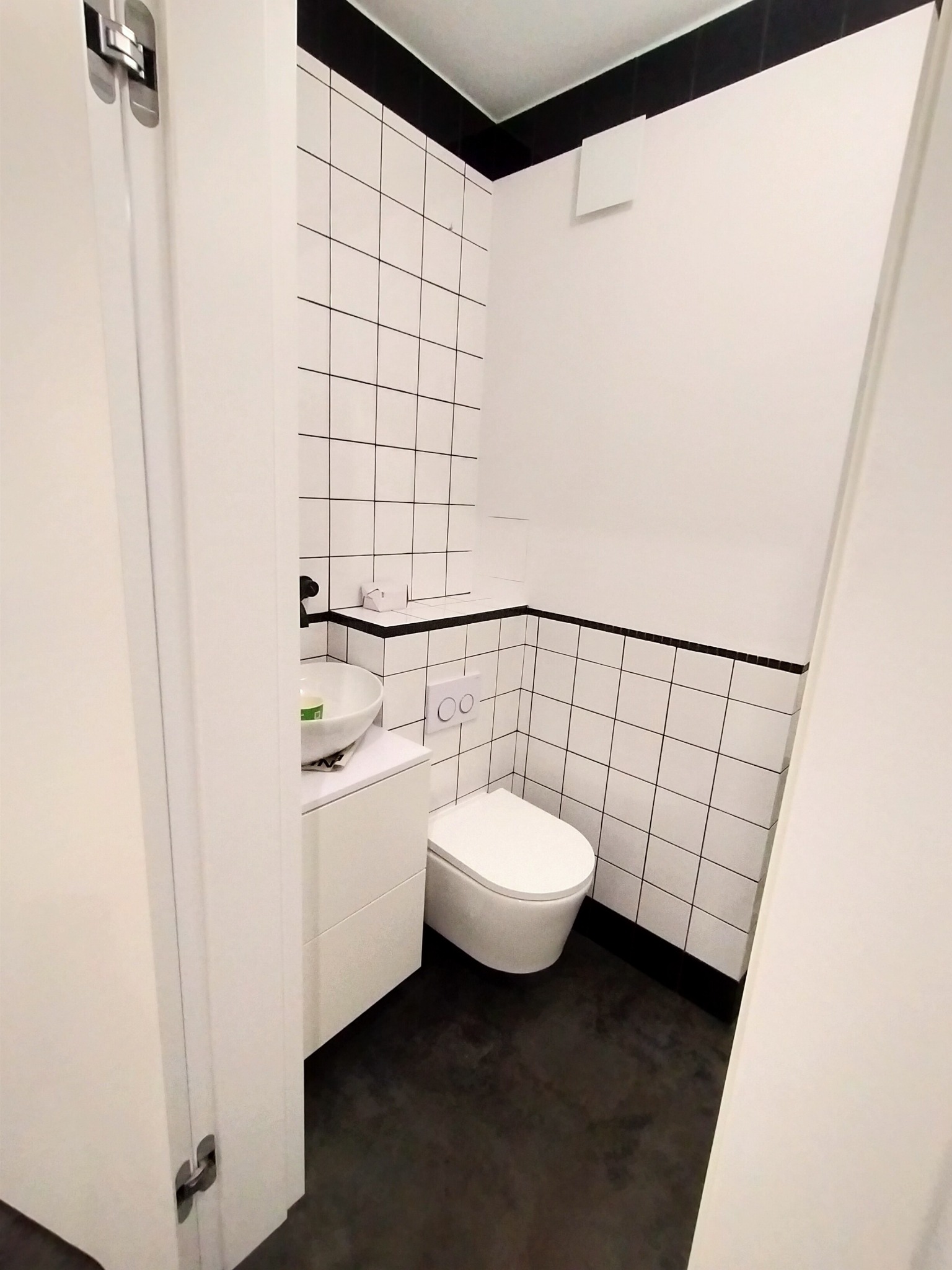 Nowoczesna łazienka z białymi kwadratowymi płytkami z czarną fugą, wiszącą miską WC, umywalką nablatową i czarną podłogą, widziana zza uchylonych białych drzwi.