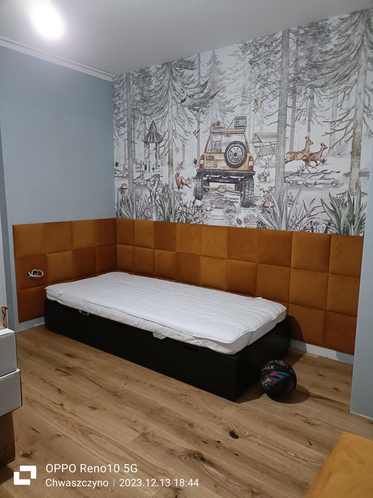 Pokój dziecięcy w Kosakowie z łóżkiem i panelem ściennym z kwadratowych, pomarańczowych poduszek, dekoracyjna tapeta w leśny motyw z samochodem terenowym w tle, na podłodze piłka.