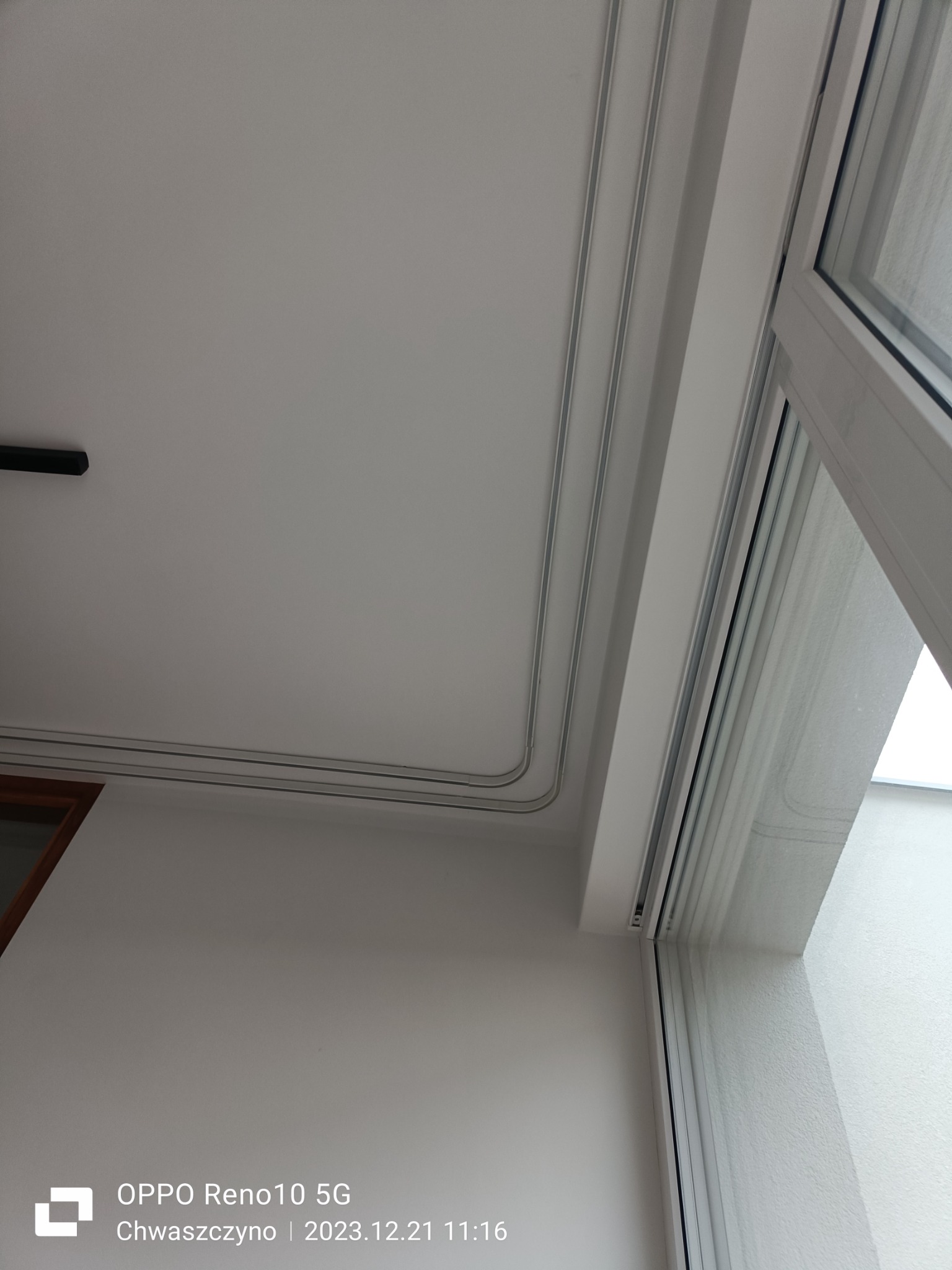 Biały sufit z widocznymi listwami karniszowymi i fragmentem okna z białymi ramami.