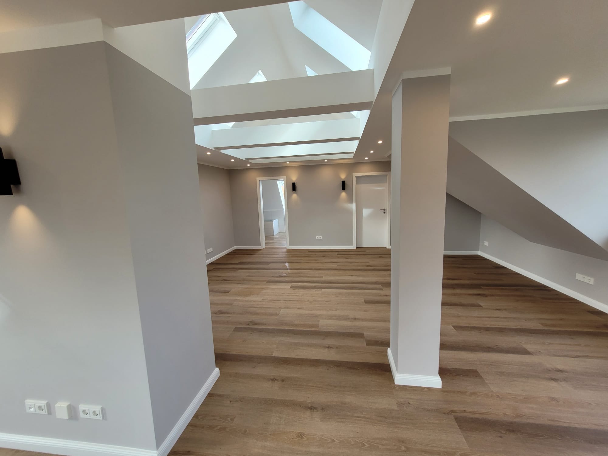 Jasne, przestronne wnętrze poddasza po remoncie z drewnianą podłogą, szarymi ścianami, białymi drzwiami i oknami dachowymi w suficie.