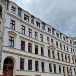 Odnowiona elewacja kamienicy z jasną fasadą, ciemnobrązową stolarką okienną i szarą dachówką, sfotografowana pod pochmurnym niebem.