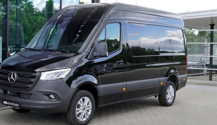 Czarny, lśniący Mercedes Sprinter z przyciemnianymi szybami, zaparkowany na zewnątrz budynku z przeszkloną fasadą.